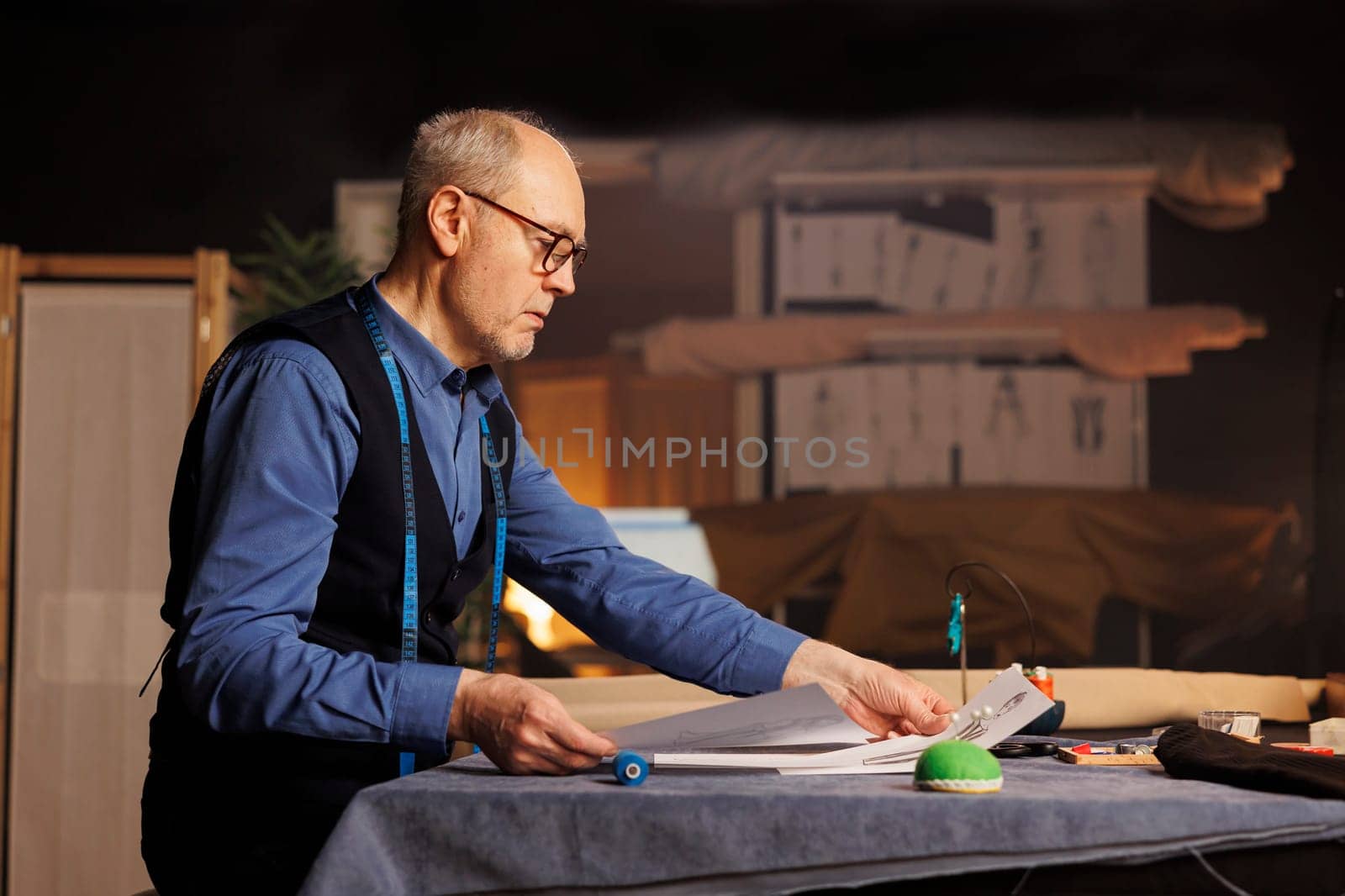 Couturer doing needlework in atelier shop workspace, checking bespoken clothing drafts. Suitmaker looking over sketch drawings, preparing to manufacture sartorial garments for client