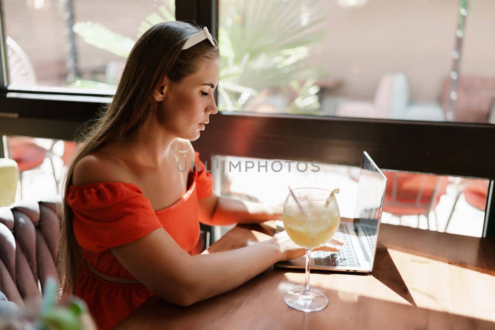 Woman works on laptop in a sunny cafe, glass of juice nearby. The setting is modern, with natural light illuminating the space. The image captures a moment of focused work