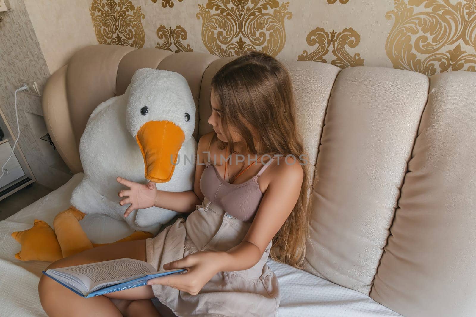 A girl is reading a book while sitting on a bed with a stuffed duck. The room has a gold and white color scheme, and there are two lamps in the room