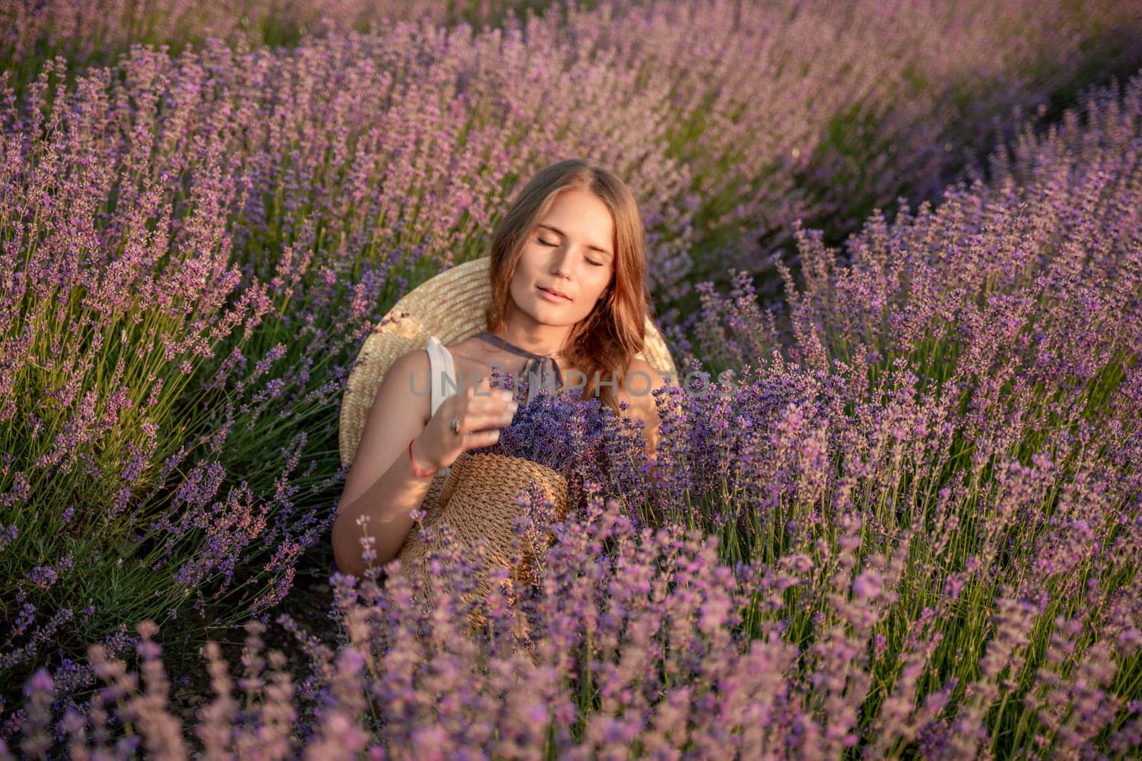 A woman is sitting in a field of lavender flowers. She is wearing a straw hat and holding a basket of flowers. The scene is peaceful and serene, with the woman enjoying the beauty of the flowers