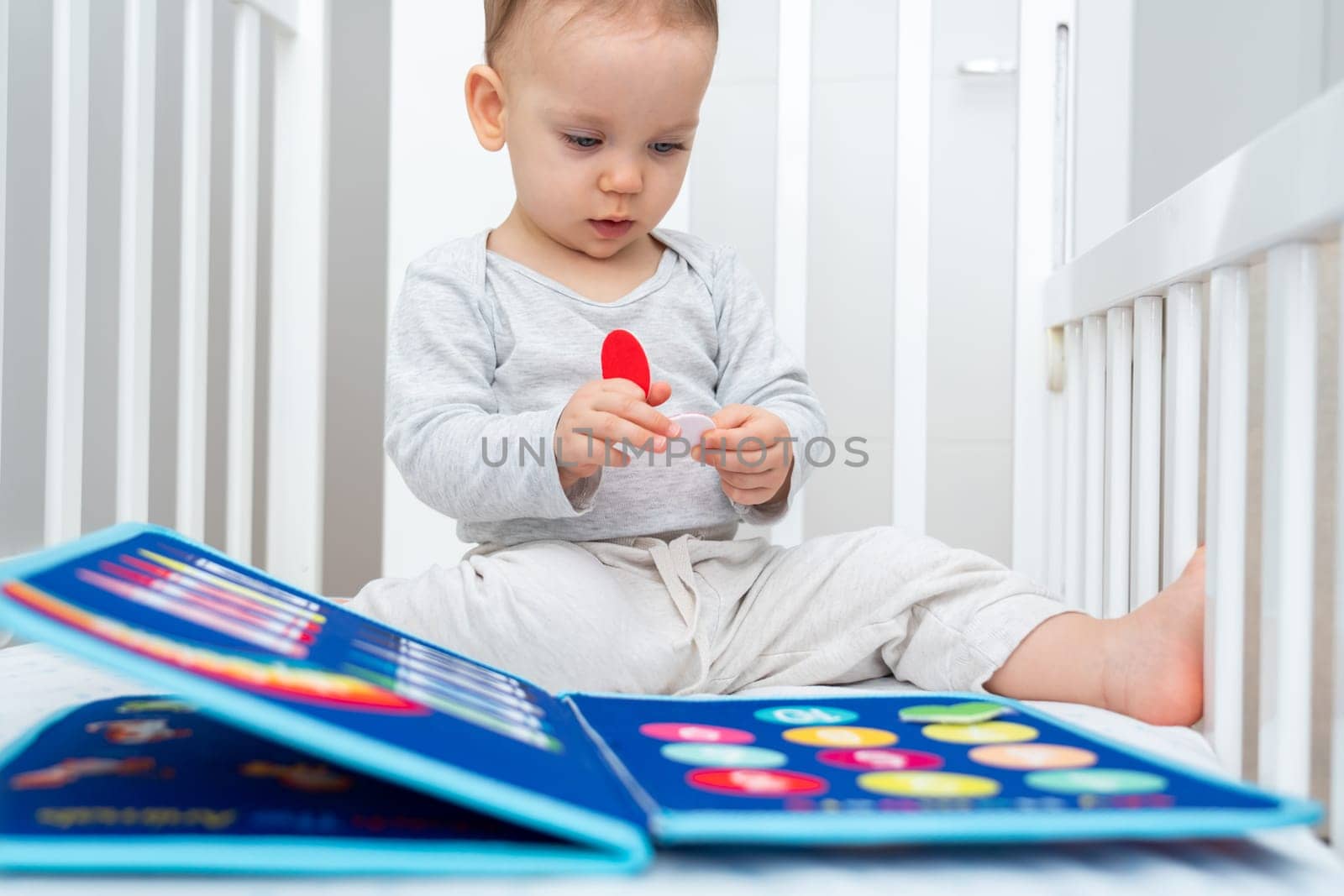 Little baby girl playing with busy book while sitting in crib. Concept of quiet books and modern educational toys by Mariakray