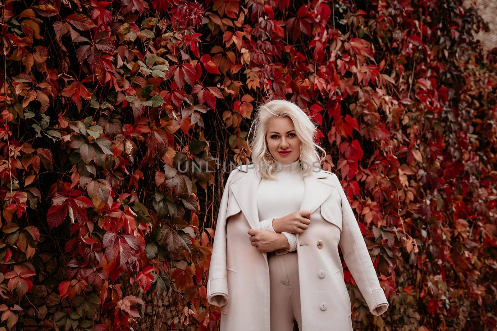 A blonde woman is standing in front of a wall covered in red leaves. She is wearing a white coat and holding her arms out to the side. Concept of warmth and comfort. by Matiunina