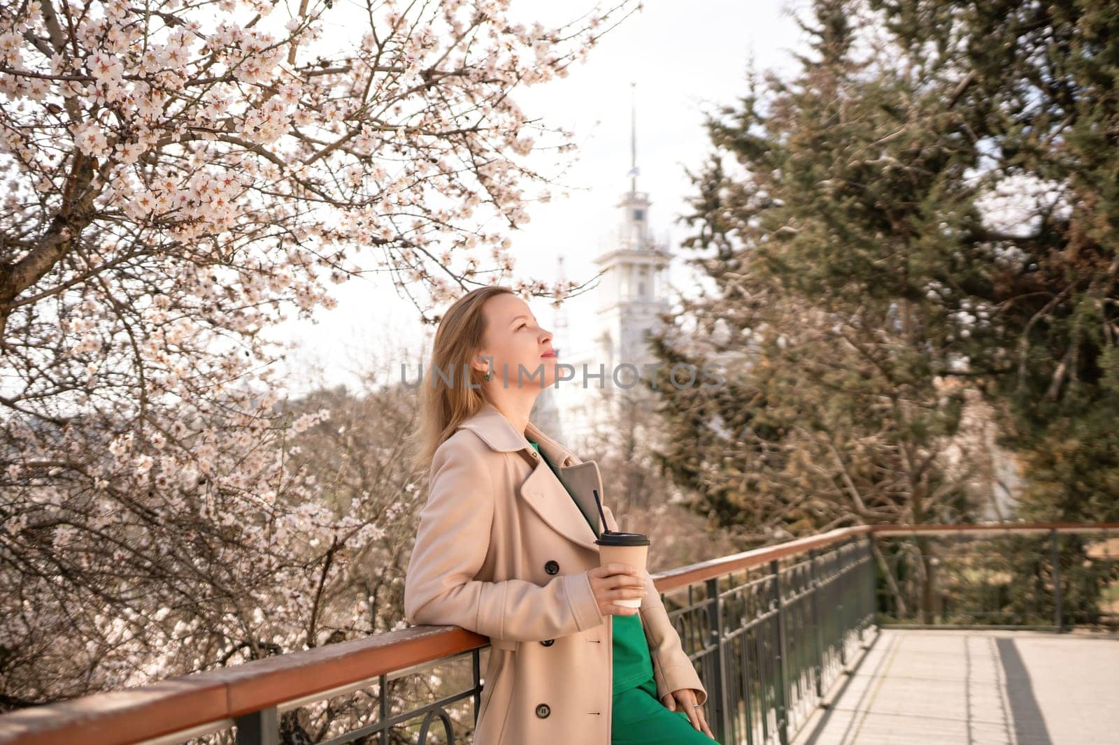 A woman is standing on a ledge with a cup of coffee in her hand. She is wearing sunglasses and a tan coat. The scene is set in a park with cherry blossoms in bloom. The woman is enjoying the view