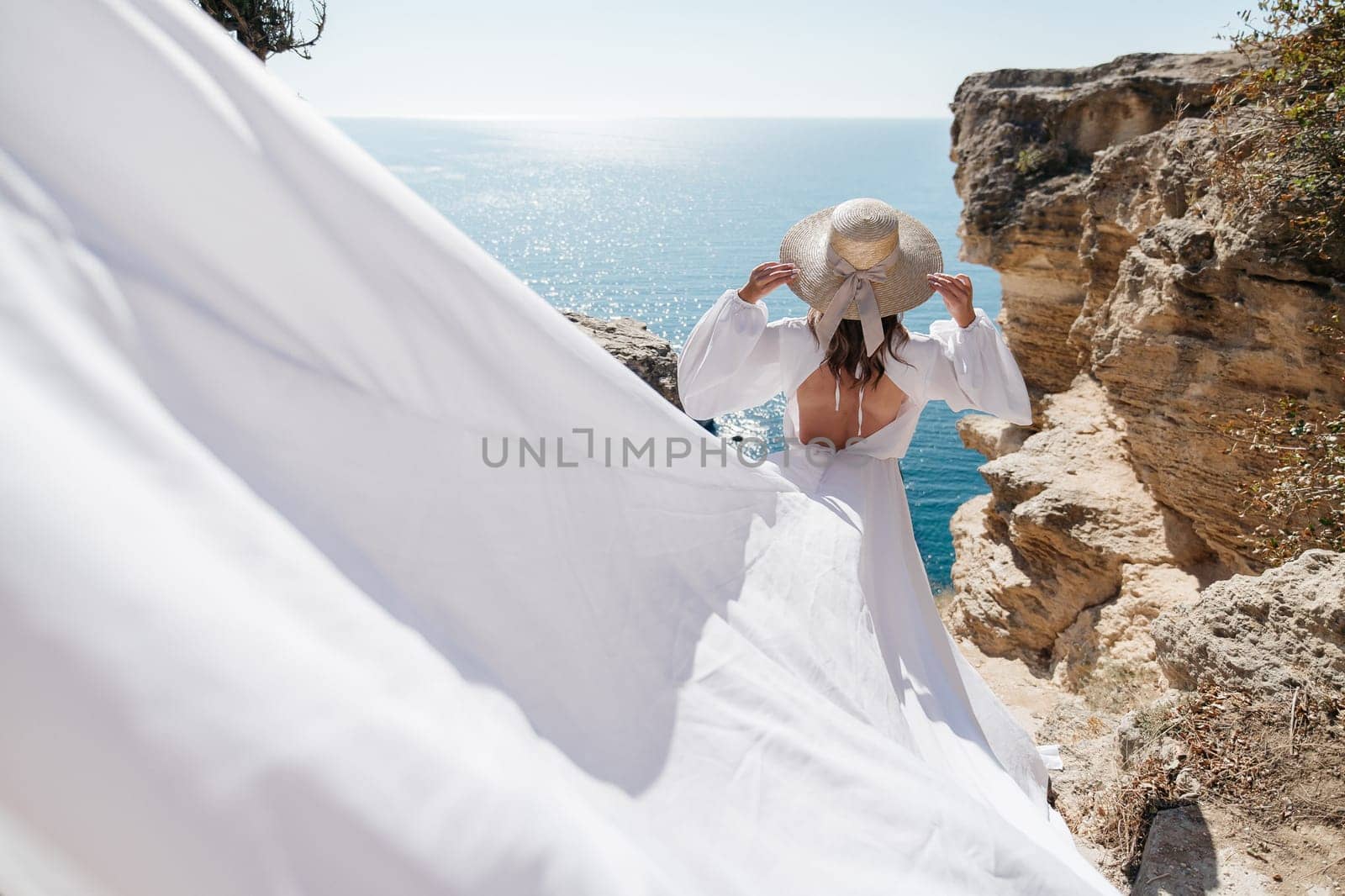 A woman in a white dress is standing on a rocky cliff overlooking the ocean. She is wearing a straw hat and she is enjoying the view