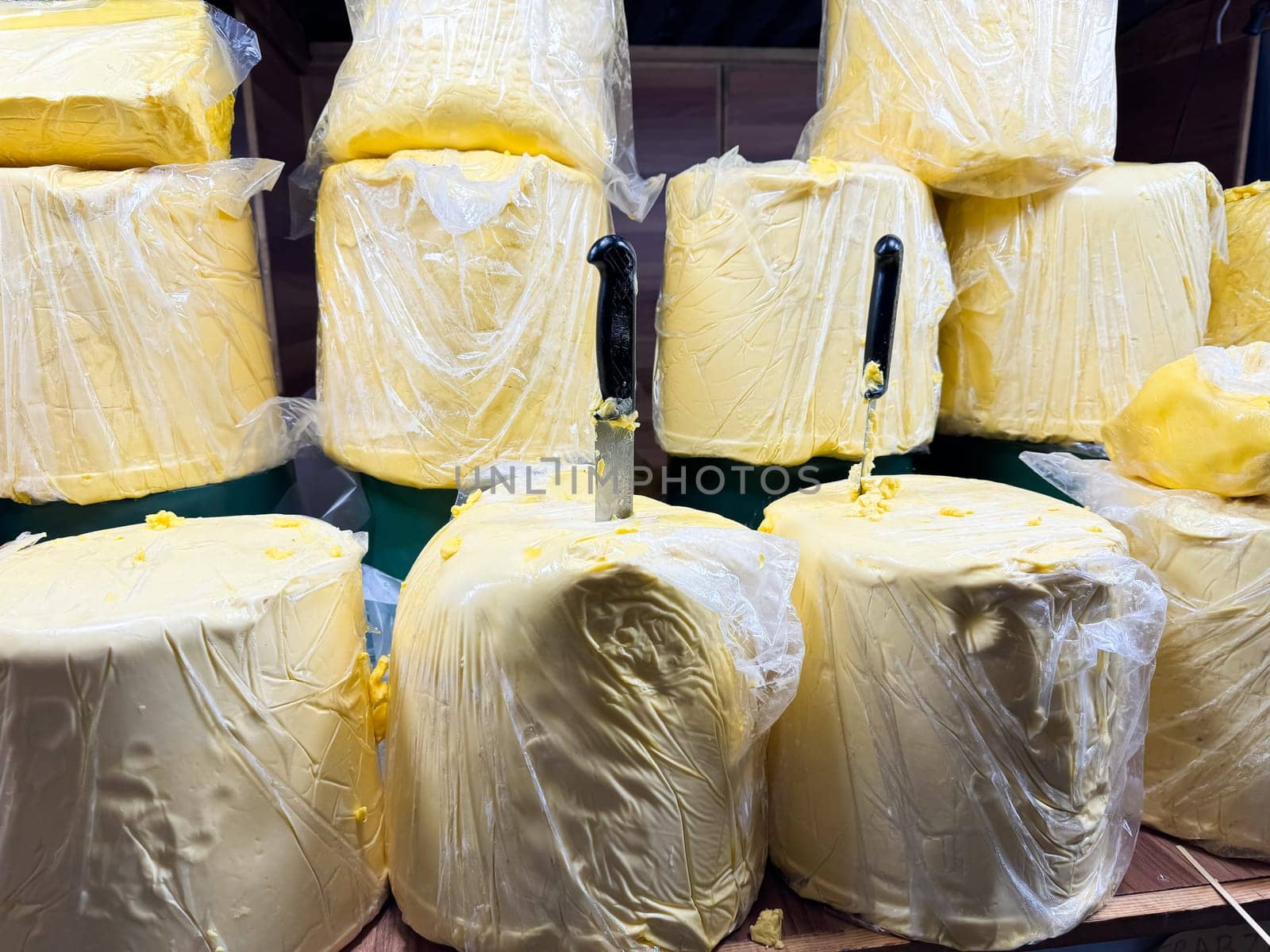 Large blocks of butter wrapped in plastic packet with knives inserted, displayed on wooden shelf at market stall. Food industry. High quality photo