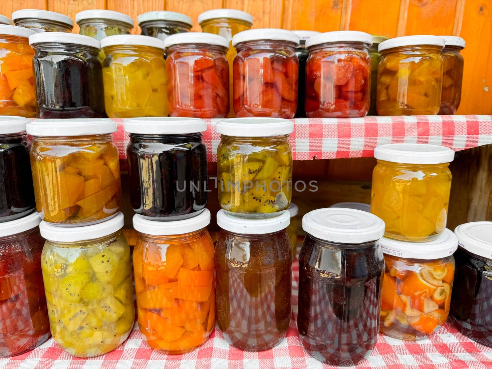 Assortment of homemade preserves in glass jars with checkered cloth, various pickled fruits and vegetables, colorful display, canned goods collection, close up with selective focus. by Lunnica