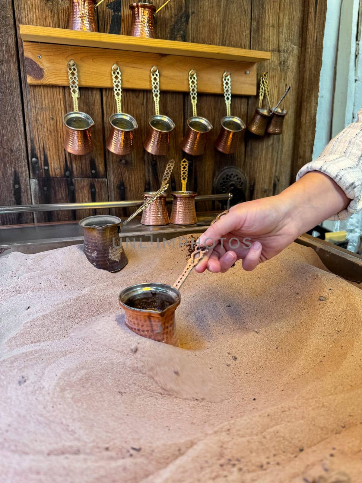 Preparing Turkish coffee in traditional copper pot on sandy stove, with hand holding the handle, copper cezves and wooden wall rack in the background. Turkish coffee culture concept. High quality