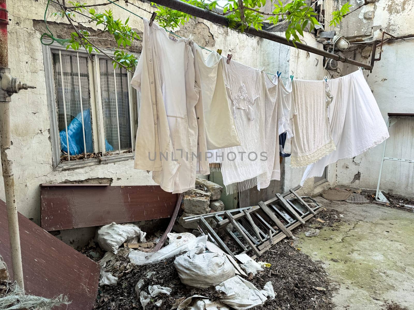 Laundry hangs on line in dilapidated city yard with overgrown leaves, broken furniture and trash, depicting neglect and everyday life and poverty in run down neighborhood. by Lunnica