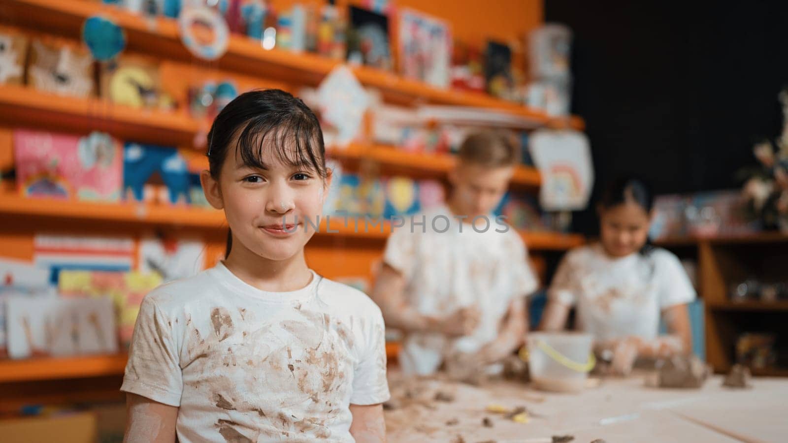 Asian highschool girl look at camera and dirty hand while diverse children modeling clay behind. Happy cute student wearing shirt while looking at camera at workshop. Blurring background. Edification.