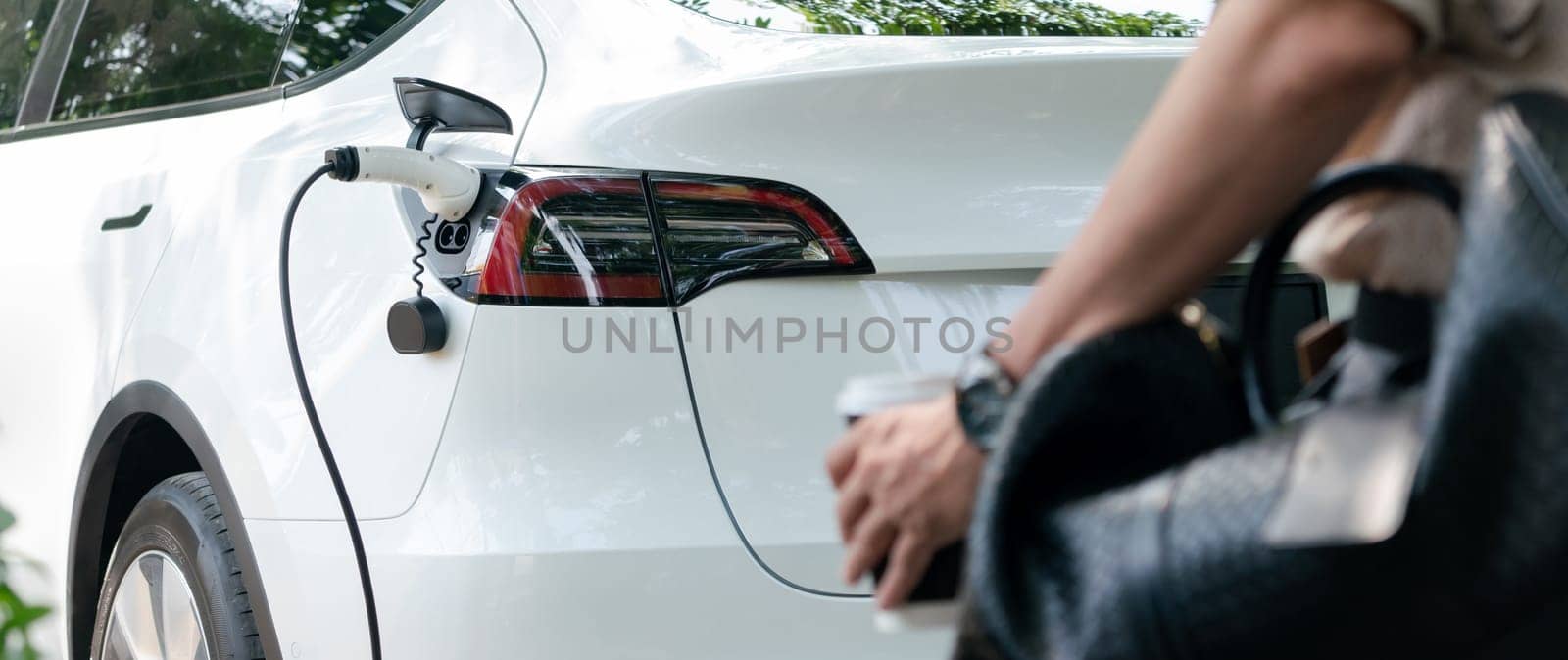 Young man recharge electric car's battery from charging station in outdoor green city park. Rechargeable EV car for sustainable environmental friendly urban travel. Panorama Expedient