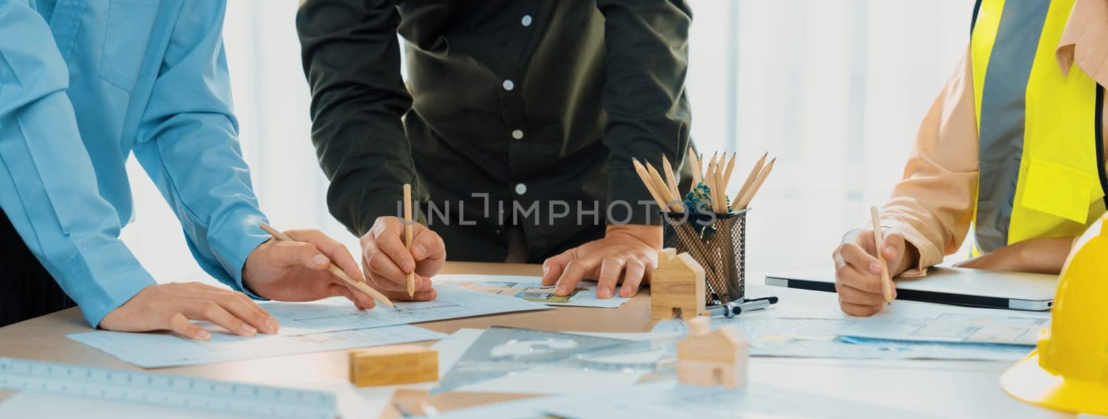 Professional architect team and safety engineer with safety helmet brainstorming about architectural project on meeting table with blueprint scatter around. Close up. Focus on hand. Delineation.