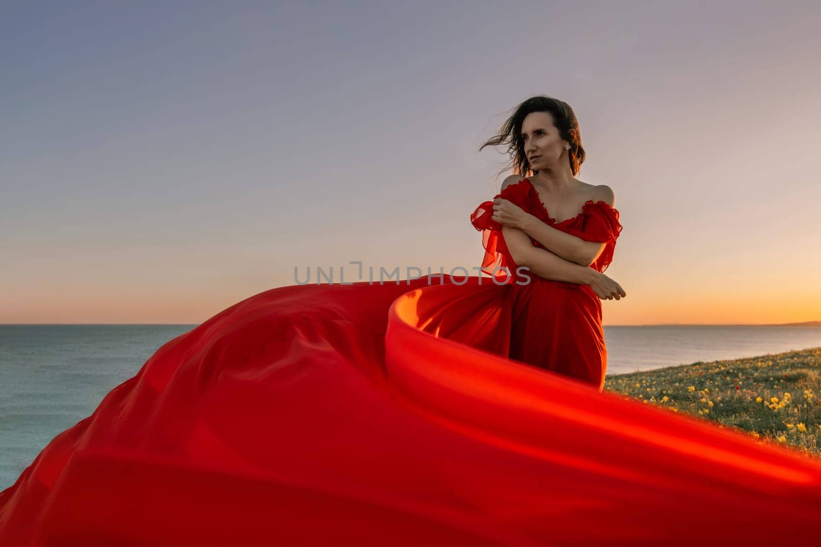 A woman in a red dress is standing on a grassy hillside. The sun is setting in the background, casting a warm glow over the scene. The woman is enjoying the beautiful view and the peaceful atmosphere. by Matiunina