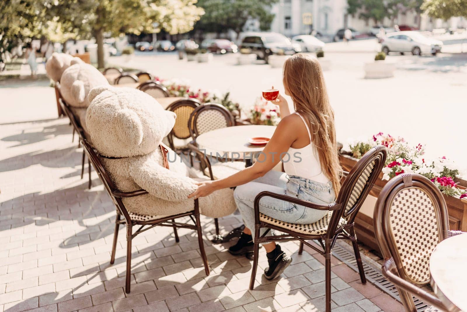 A woman sits cafe with a teddy bear next to her. The scene is set in a city with several chairs and tables around her. The woman is enjoying her time at the outdoor cafe