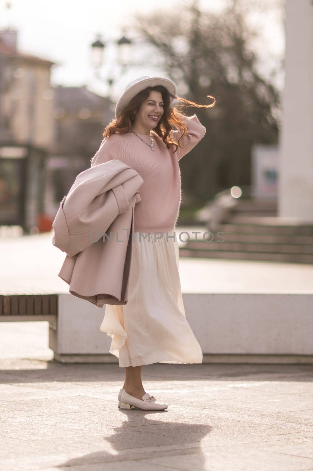 A woman wearing a pink sweater and white skirt is smiling and holding a brown bag. The scene has a warm and cheerful mood, with the woman looking happy and confident