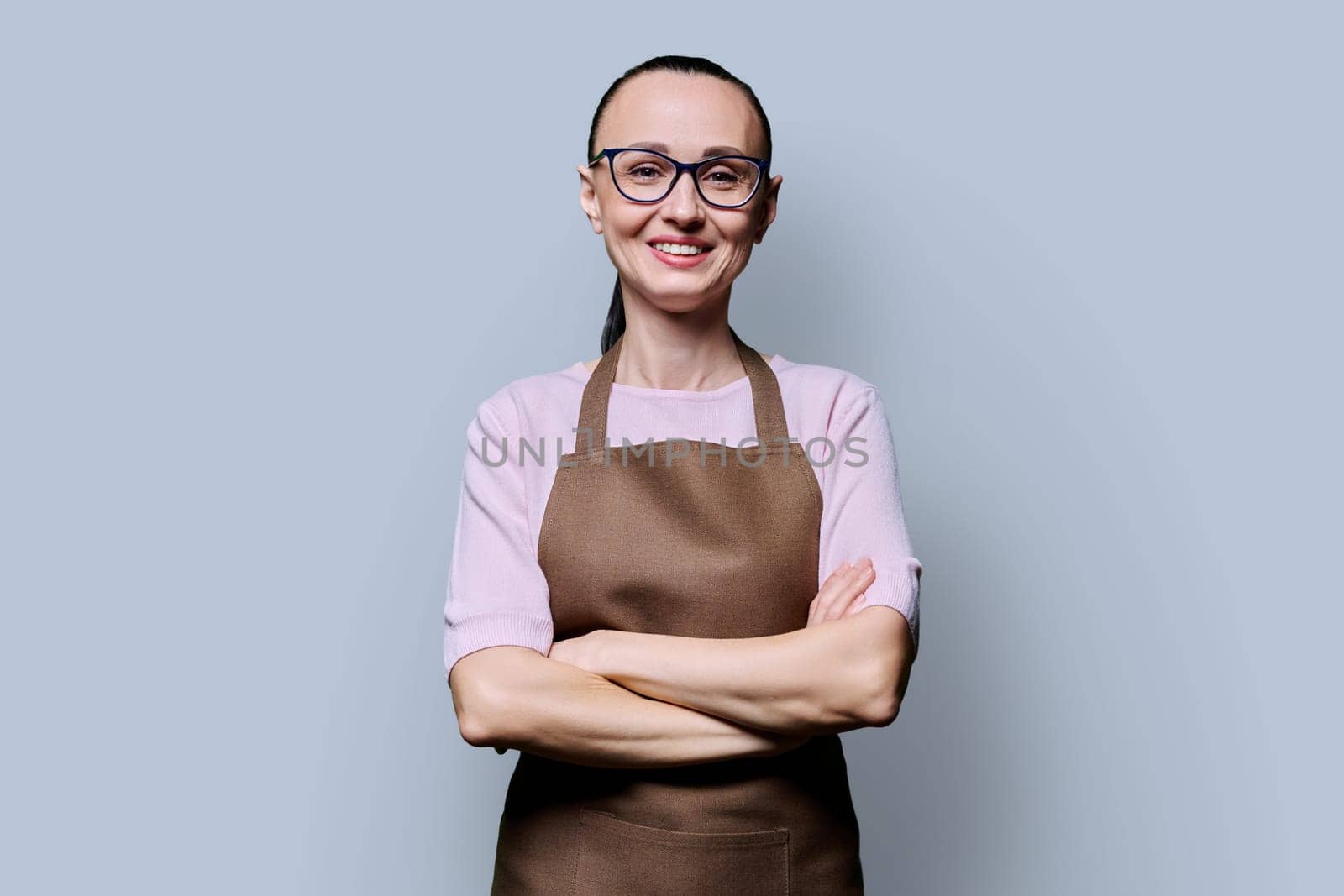 Portrait of confident smiling 30s woman in apron on grey background. Successful female small business owner, service worker looking at camera with crossed arms. Staff, management, advertising, people