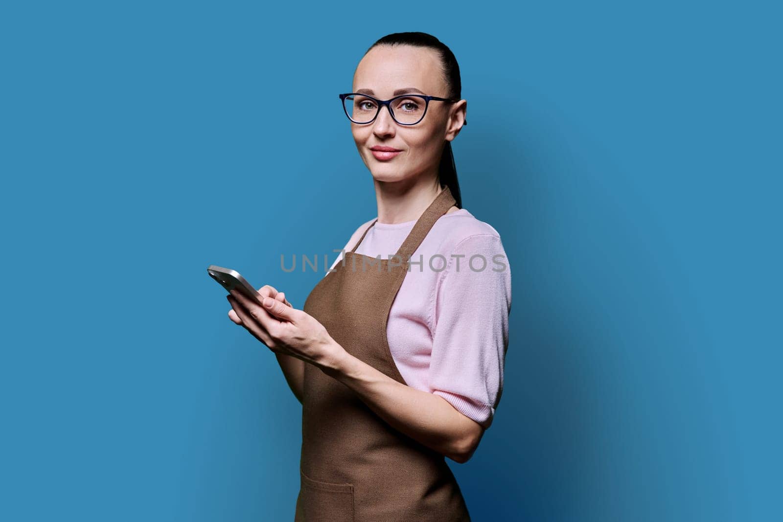 Portrait of 30s woman in apron with smartphone looking at camera on blue background by VH-studio