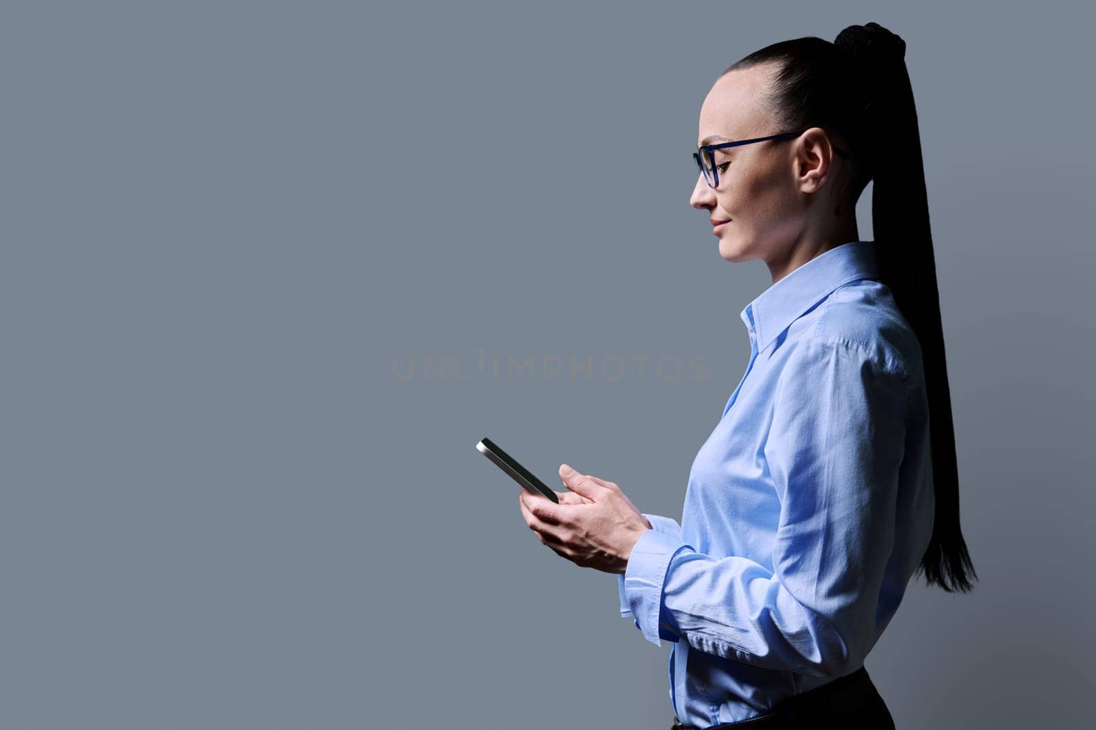 Profile view 30s smiling woman using smartphone on grey background. Middle aged female looking at phone in hands. Technologies mobile apps applications internet work business leisure communication