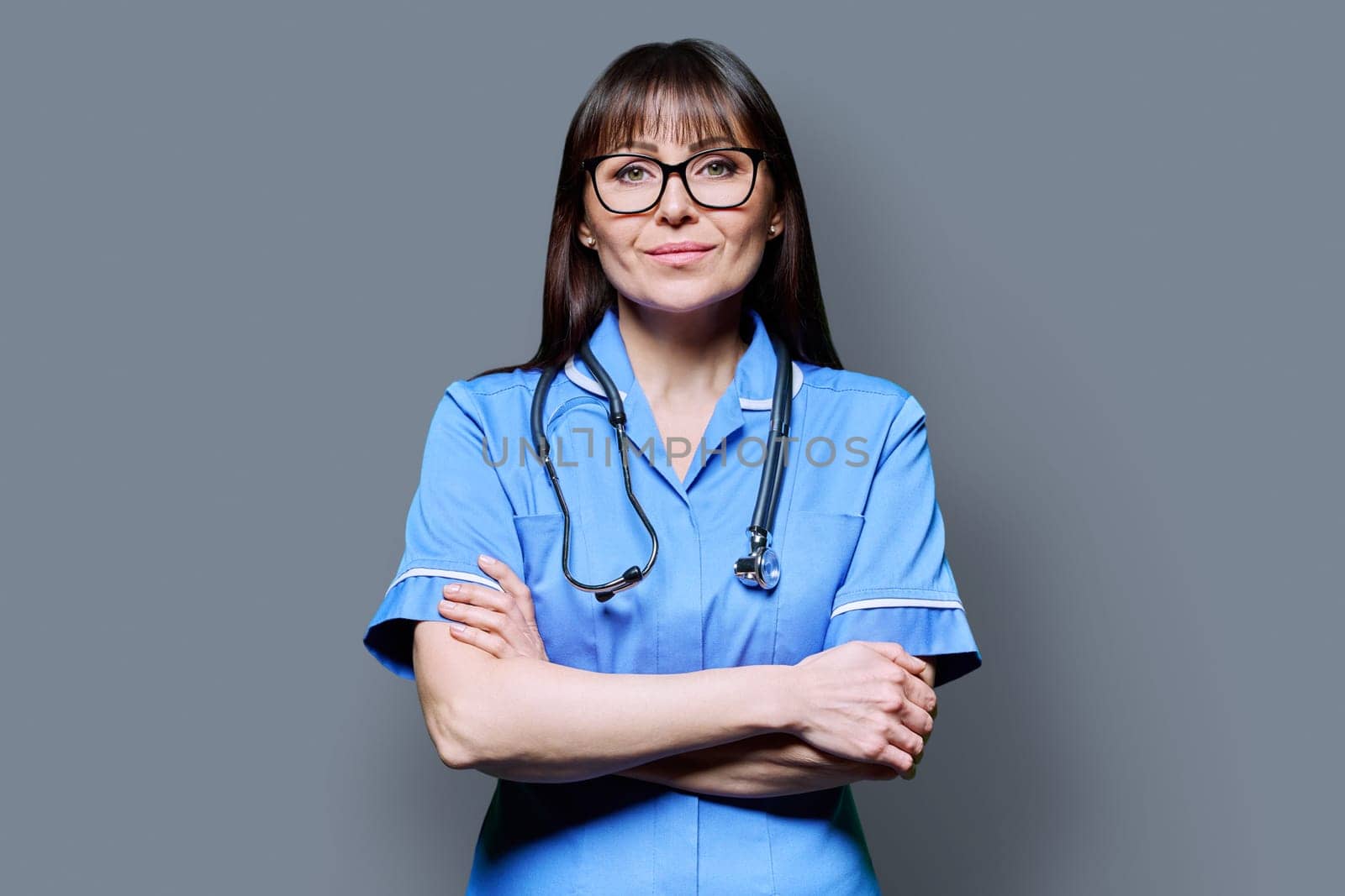 Portrait of smiling middle-aged woman nurse in blue with crossed arms with stethoscope on grey studio background. Medical services, nhs, health, professional assistance, medical care concept