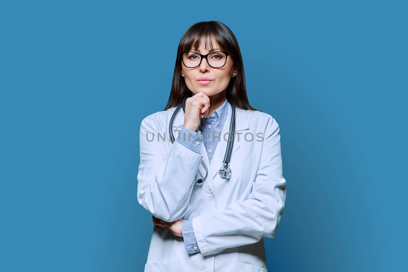 Confident serious middle-aged female doctor in white lab coat with stethoscope, looking at camera on blue studio background. Healthcare, medicine, staff, treatment, medical services concept