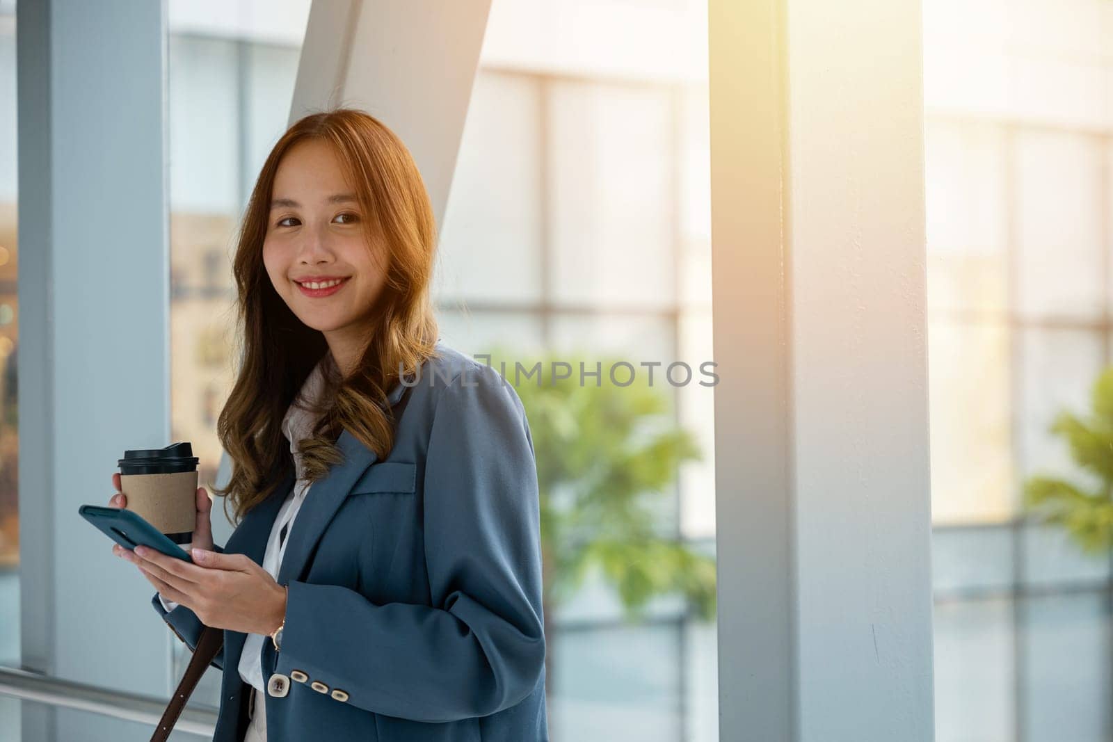 Smiling Asian woman with coffee and smartphone walking on escalator by Sorapop