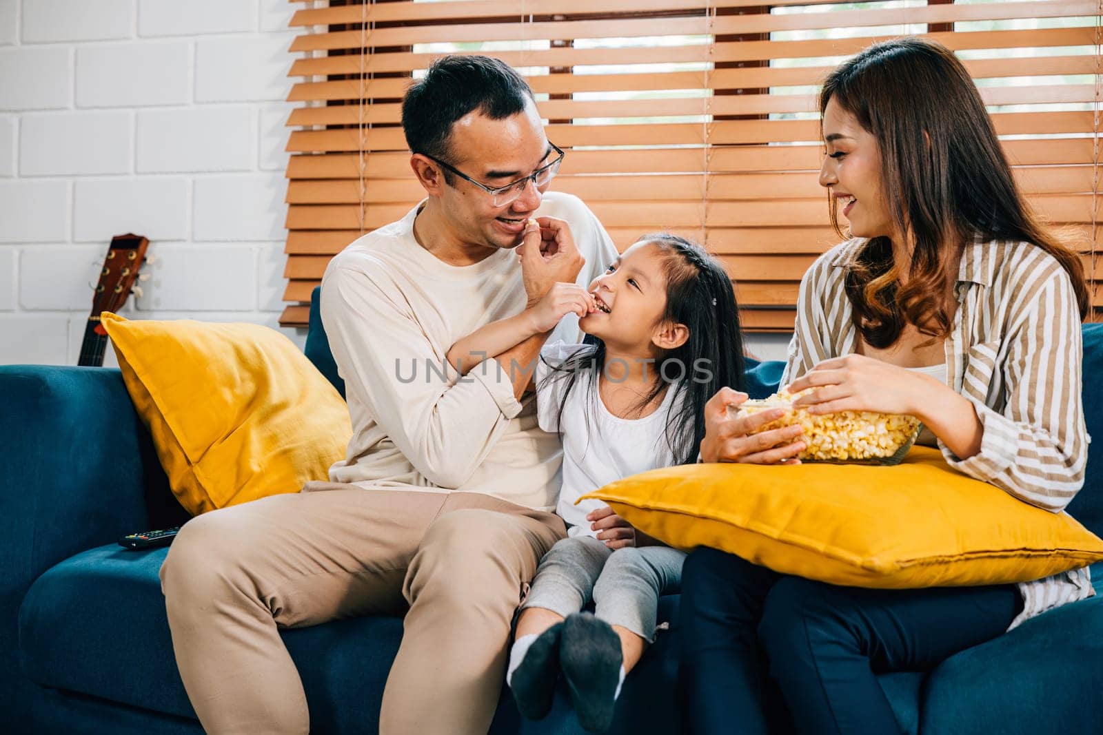 A happy family filled with joy watches TV while eating popcorn on modern sofa at home by Sorapop