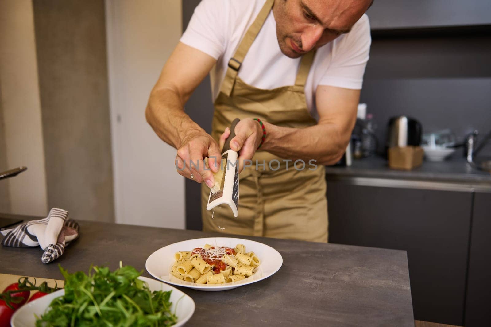 Close-up chef cooking dinner in stylish modern minimalist home kitchen interior, grating parmesan cheese over a plate with freshly boiled pasta with tomato sauce. Ingredients on the kitchen countertop