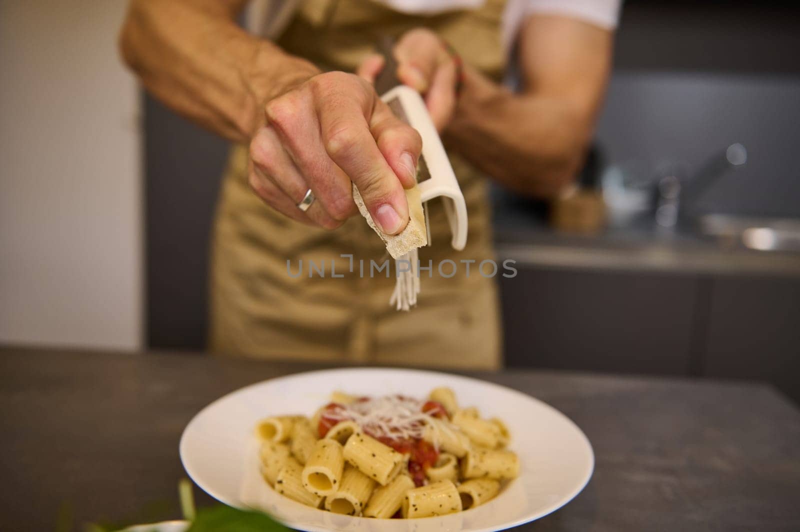 Selective focus on hands of male chef in an beige apron, grating parmesan cheese into a dish of spaghetti with tomato sauce, cooking dinner according to traditional Italian recipe in the home kitchen.