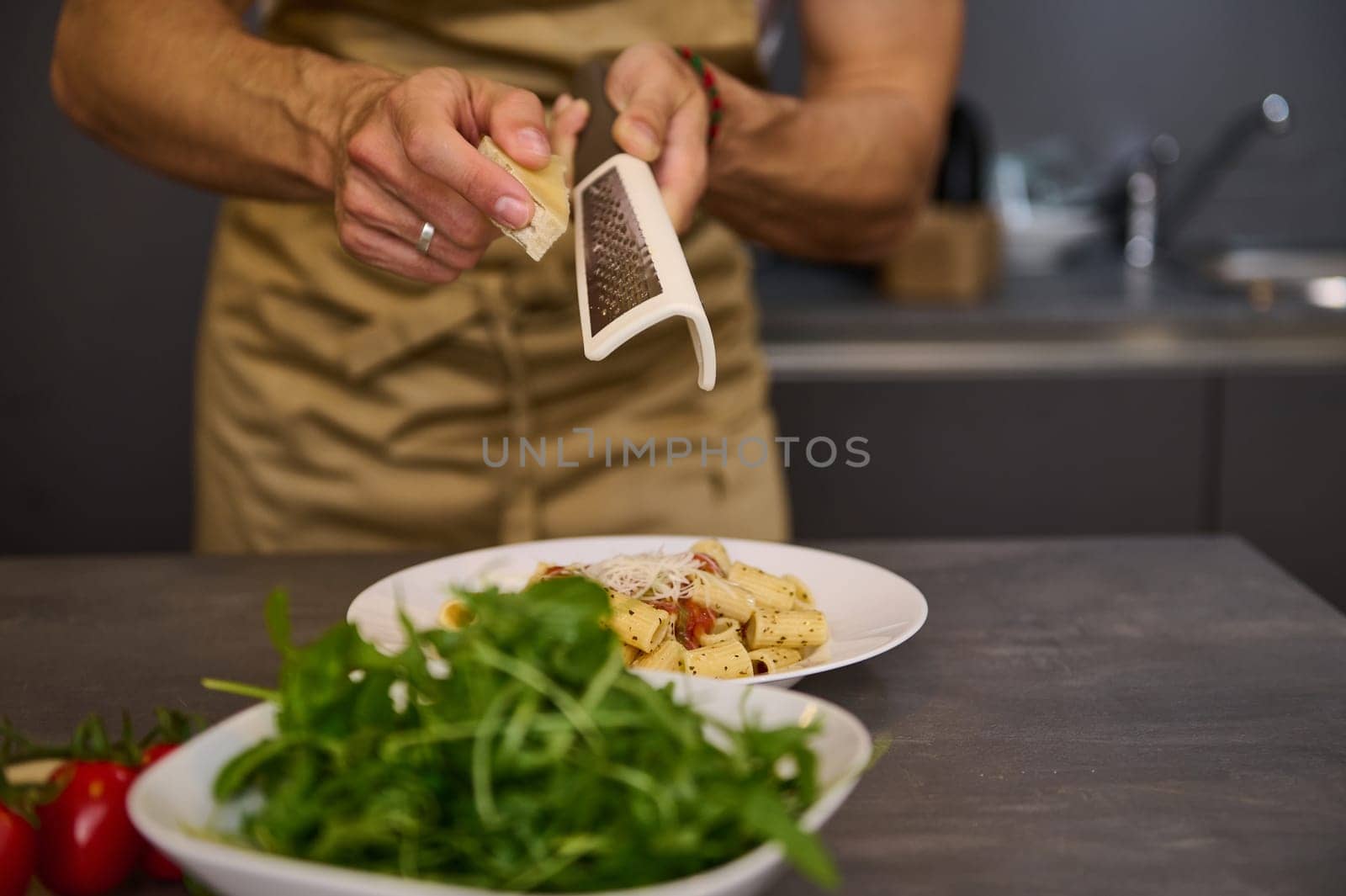 Closeup man cooking dinner at home, preparing Italian pasta with tomato sauce and pouring grated parmesan cheese. A bowl with fresh clean arugula leaves on the foreground by artgf