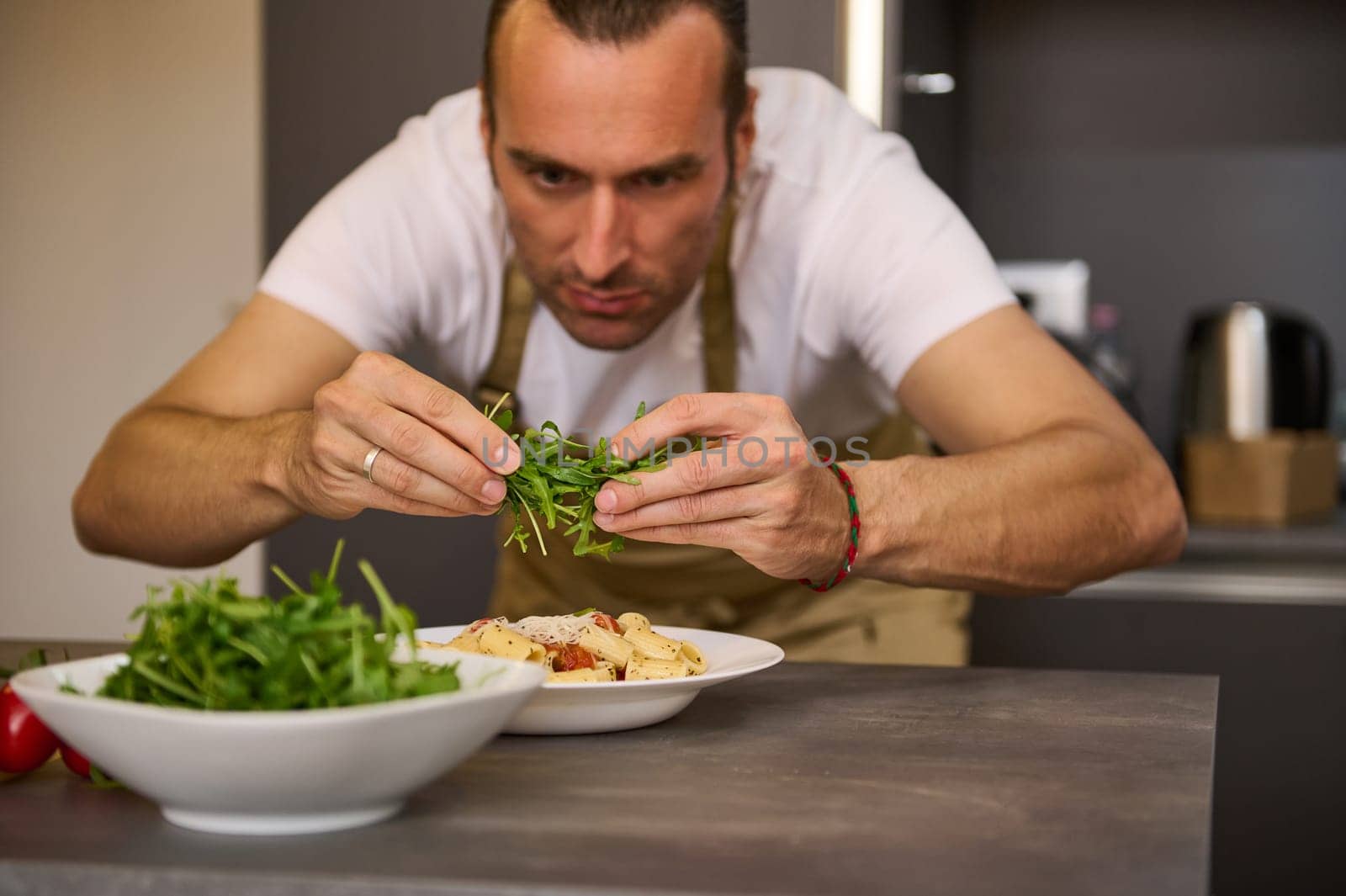 Male chef adding fresh green arugula leaves to Italian pasta with tomato sauce, garnishing the dish. A man preparing dinner in home kitchen. Selective focus on hands holding greens for seasoning food