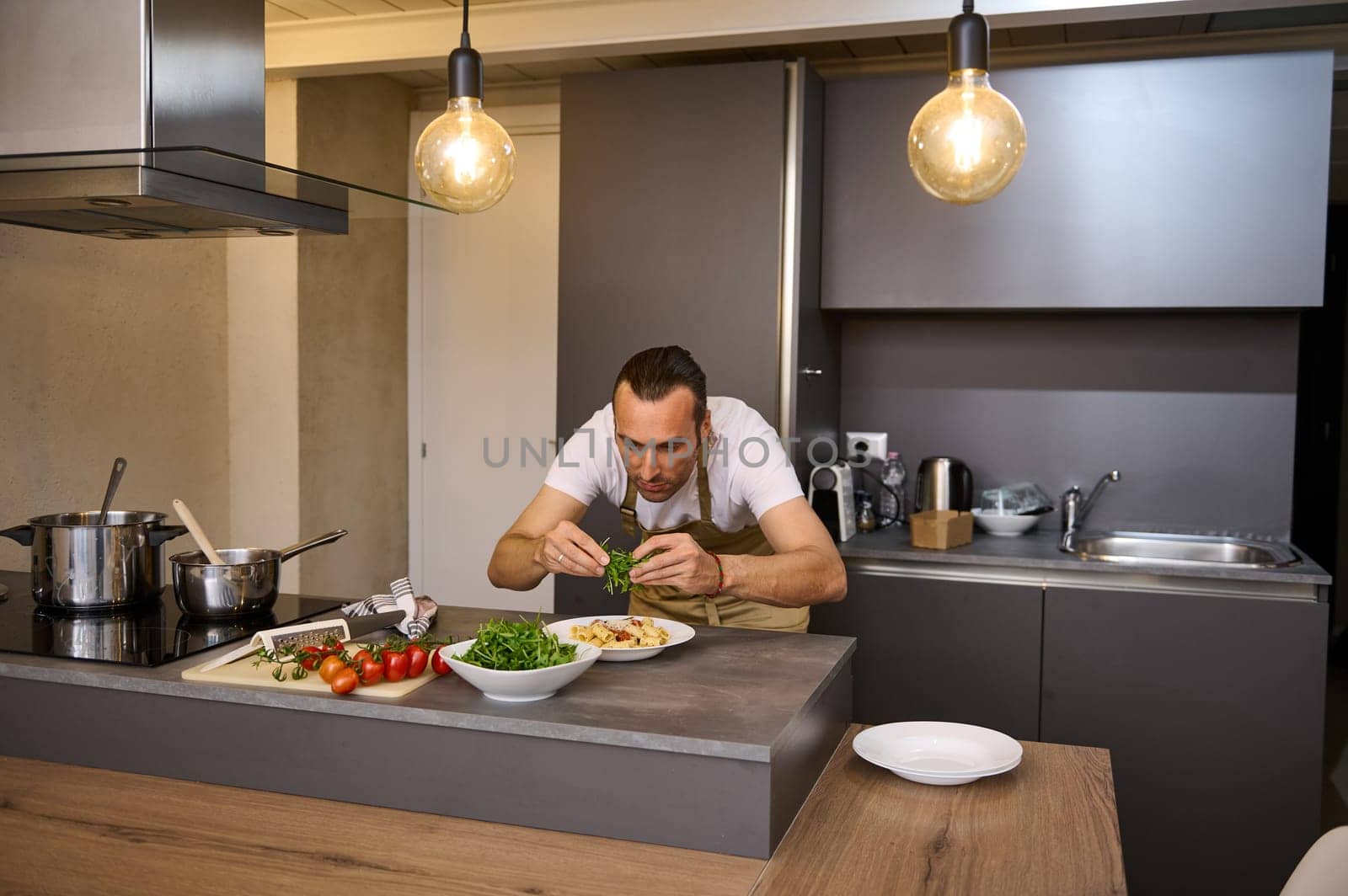 Male chef adding fresh green arugula leaves to Italian pasta with tomato sauce, garnishing the dish. A man preparing dinner in home kitchen. Selective focus on hands holding greens for seasoning food by artgf