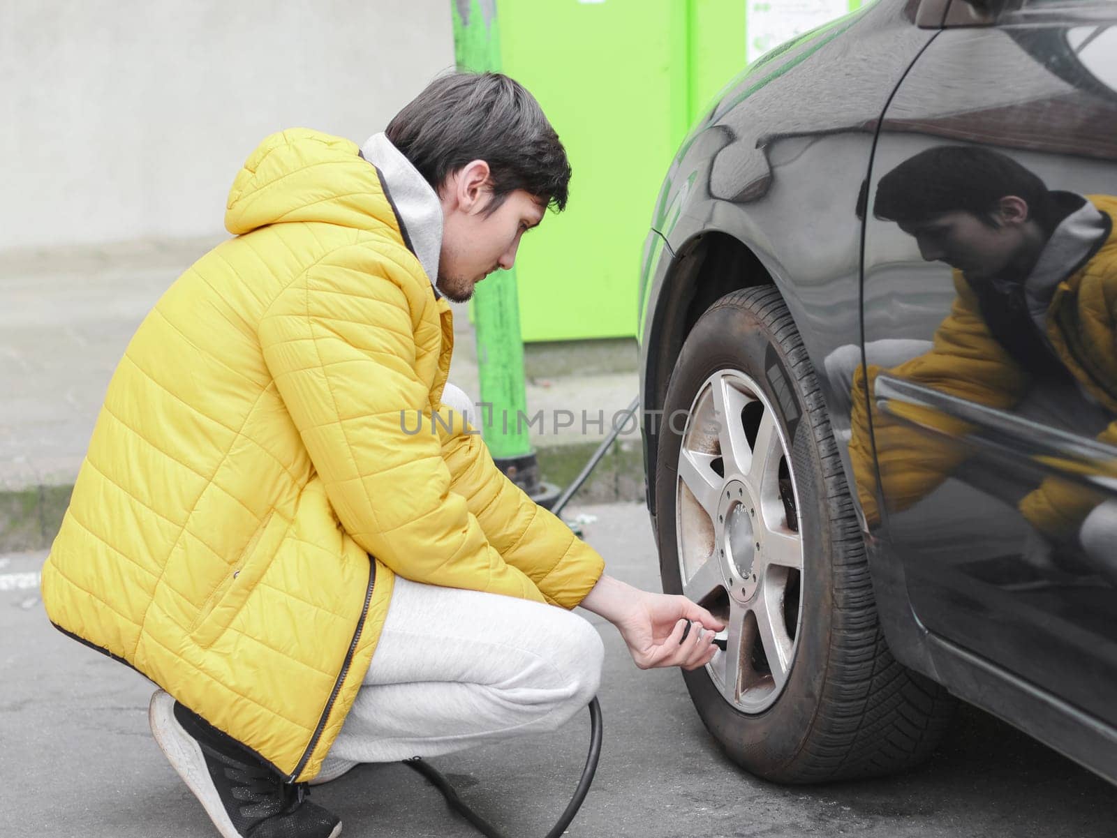 Young caucasian man in a yellow jacket and gray sweatpants, squatting, spins the bearing with his fingers into the wheel of a car for pumping air in a parking lot, side view with selective focus and closeup.Gas station concept.