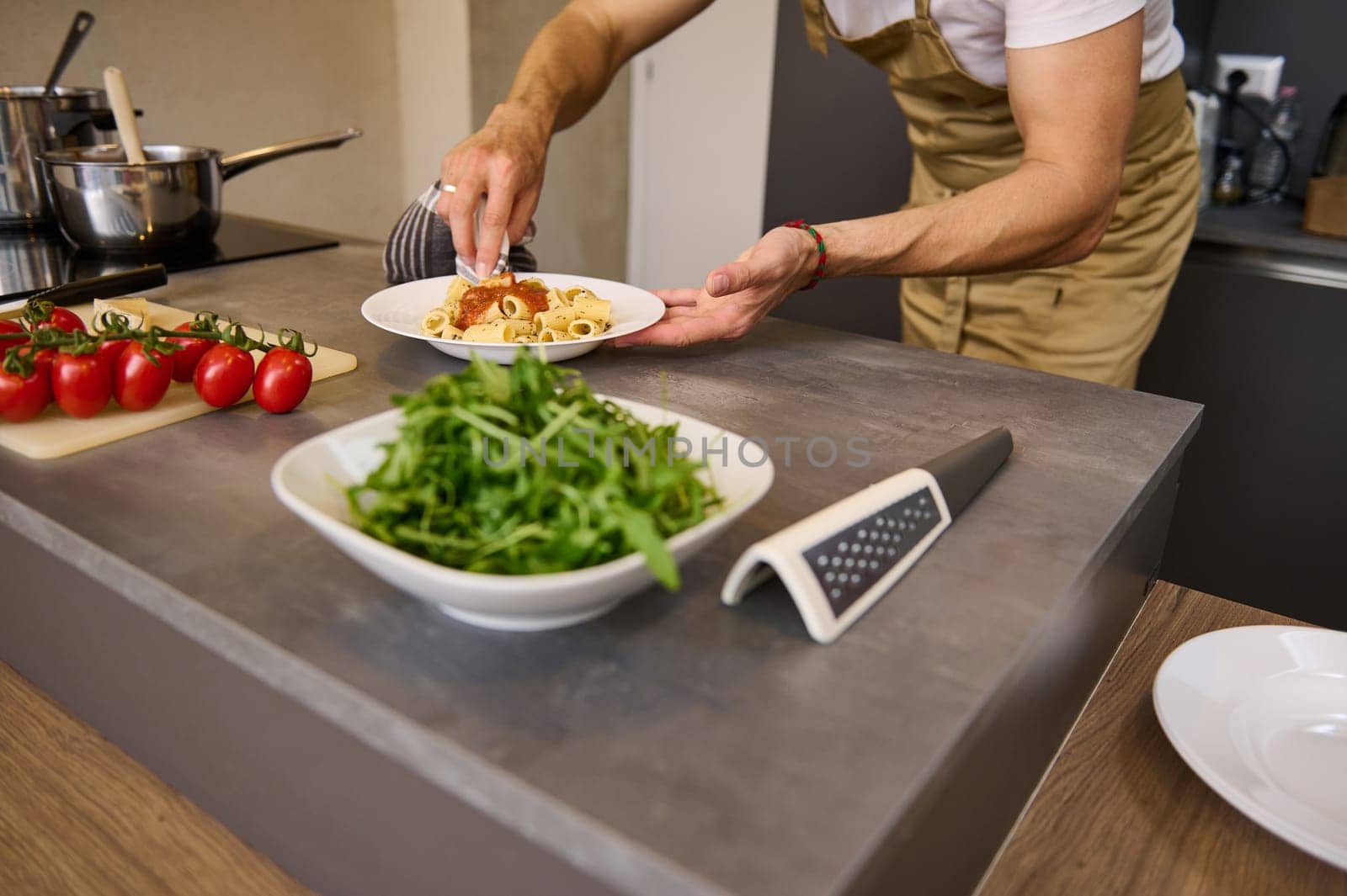 Close-up chef's hands using kitchen towel, wiping away traces of tomato sauce on a plate of freshly prepared Italian pasta. Ingredients and kitchen utensils in the kitchen table on blurred background.
