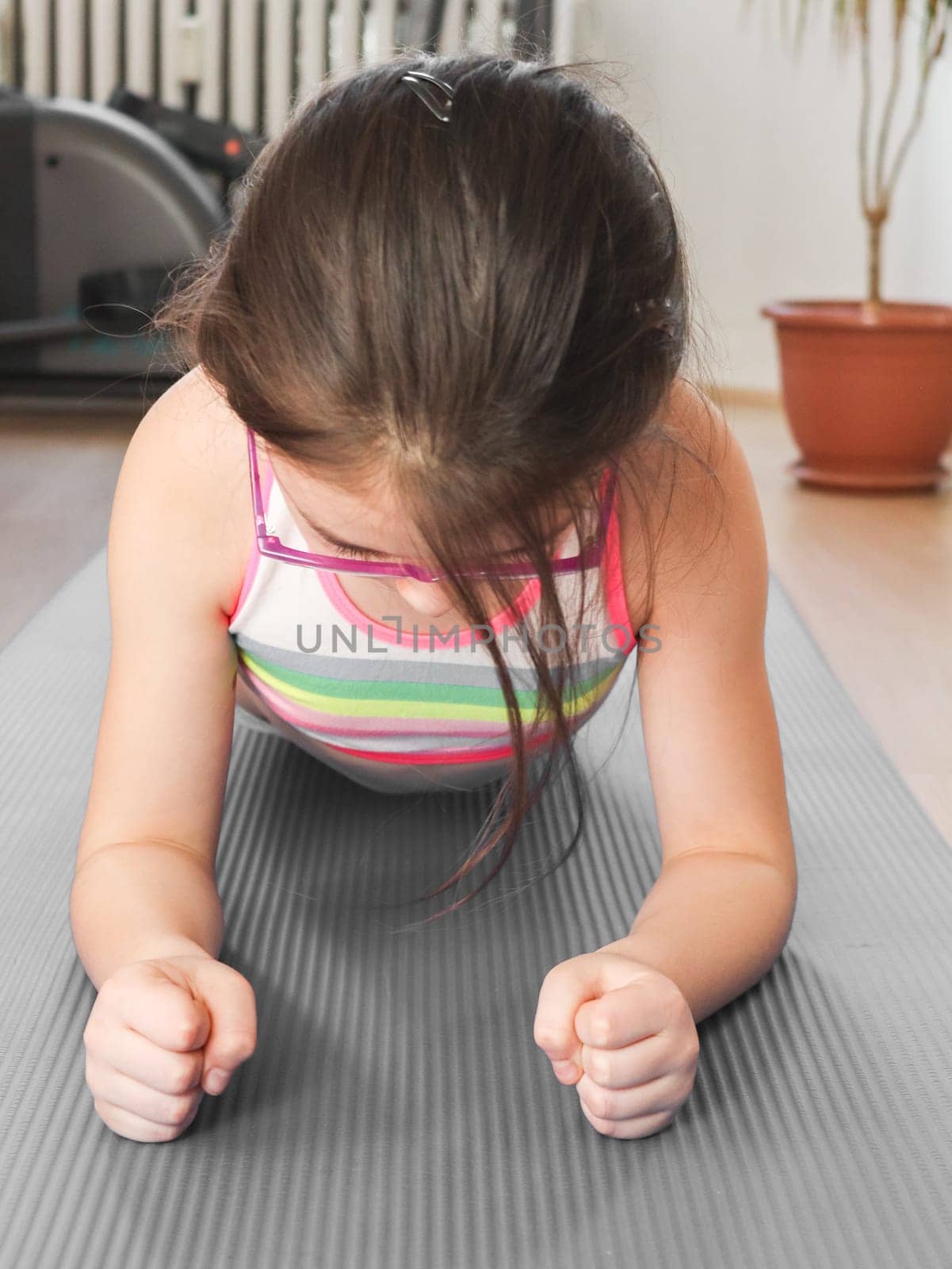 A little caucasian girl in a tracksuit and glasses does a plank exercise, clenching her hands into fists on a gray rug in a room on a wooden floor, close-up side view. The concept of home fitness, child sports and healthy lifestyle.