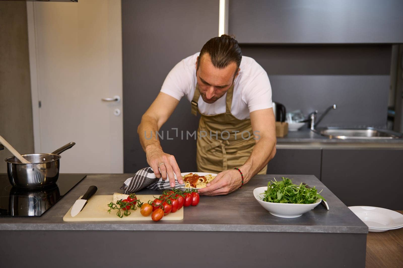 European man chef in beige apron, standing at kitchen counter and using kitchen towel, removing traces of tomato sauce on a plate of freshly prepared Italian pasta. People. Culinary. Italian cuisine