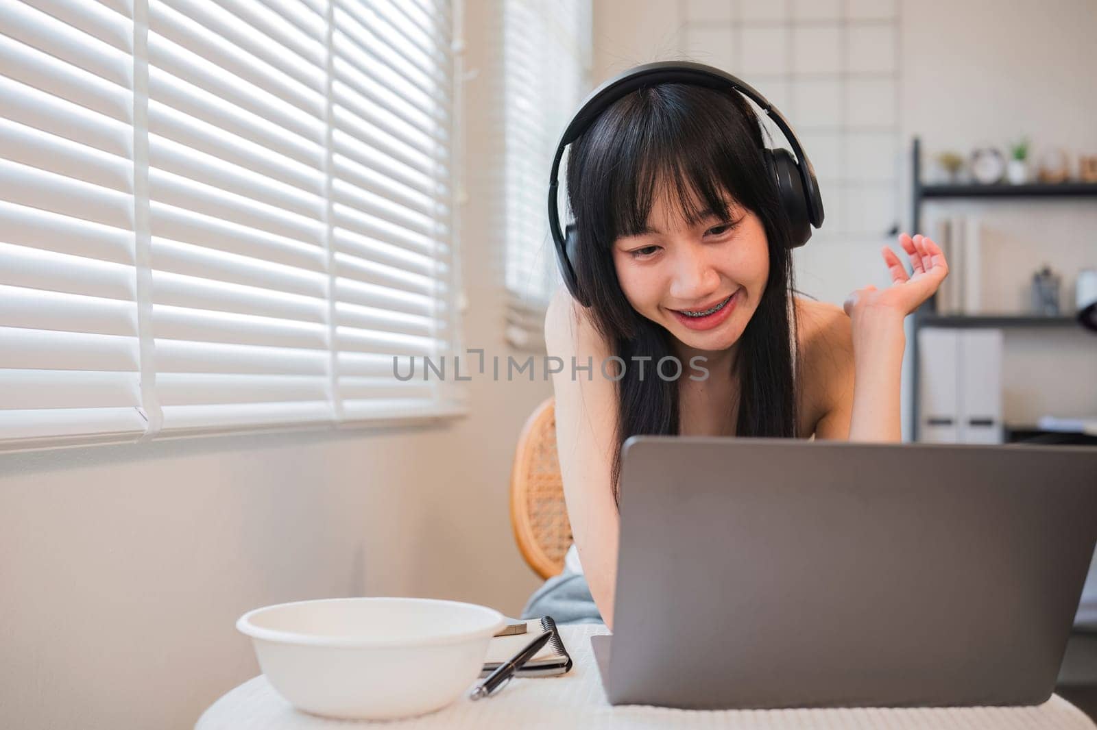 A woman is sitting at a table with a laptop and a bowl of food. She is wearing headphones and she is smiling. The scene suggests a casual and relaxed atmosphere