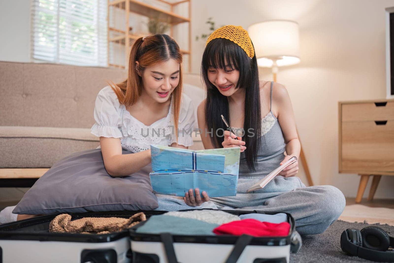 Two women are looking at a map and writing in a notebook. They are sitting on a bed with suitcases and a lamp nearby