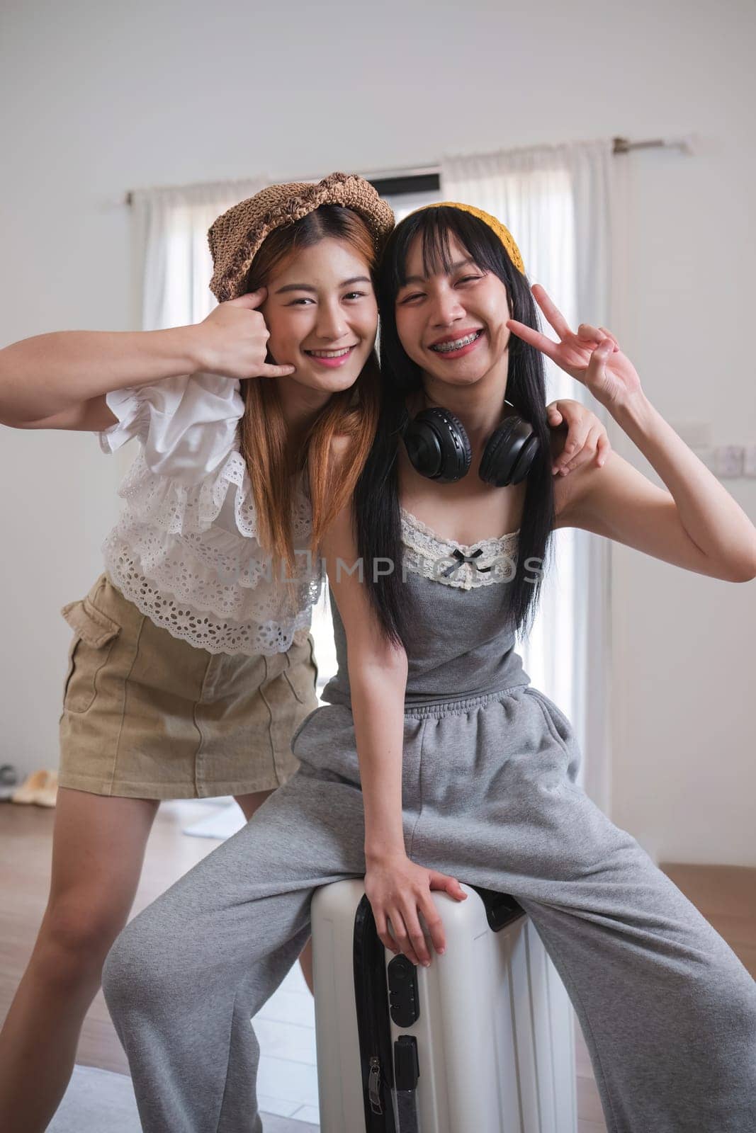 Two women are posing for a picture, one wearing a white shirt and the other wearing a gray outfit. They are both smiling and one of them is holding a suitcase. Scene is lighthearted and fun