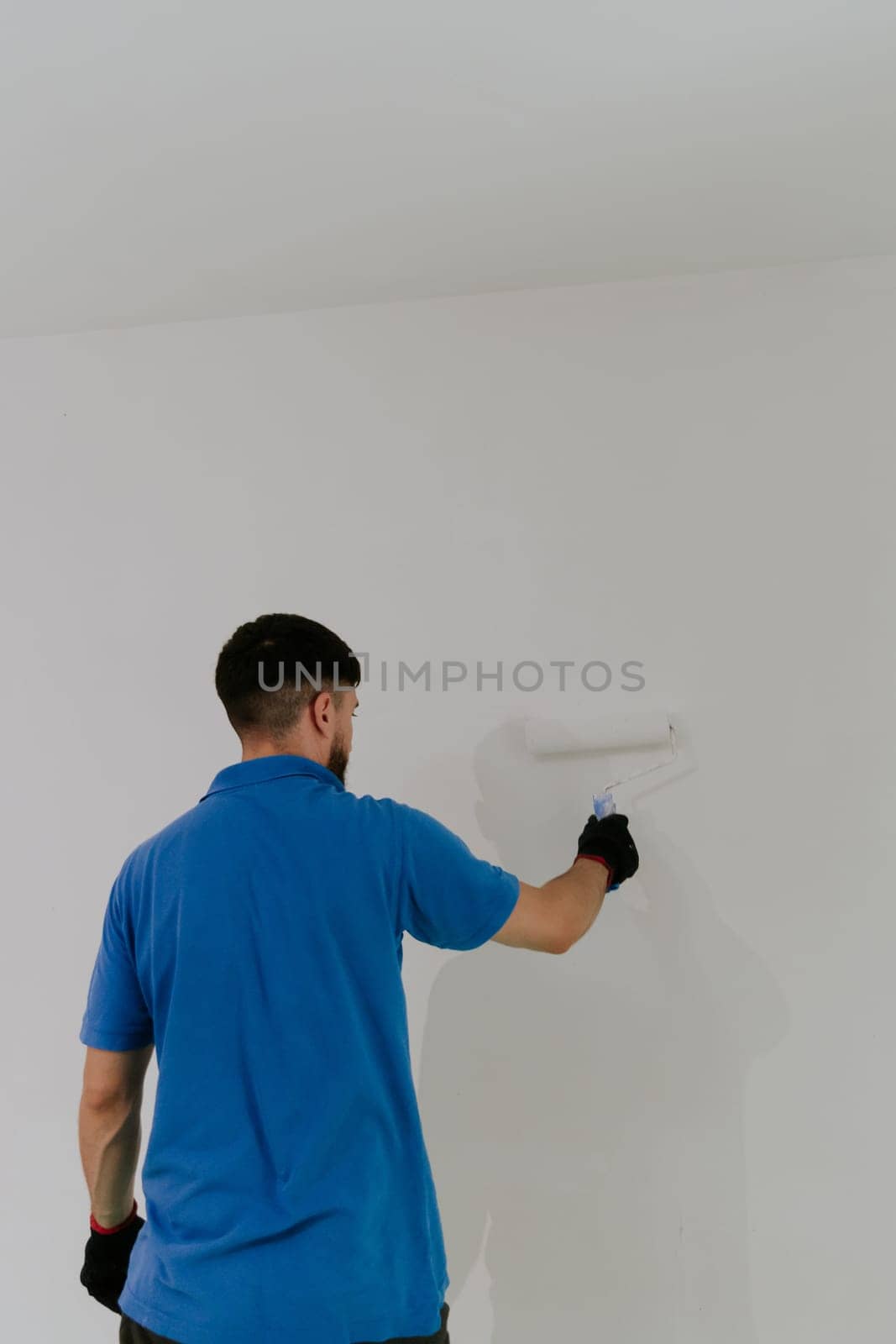 One young handsome dark-haired bearded Caucasian man in a blue t-shirt stands with his back and paints a wall with a roller of white paint, bottom side close-up view with selective focus and copy space.