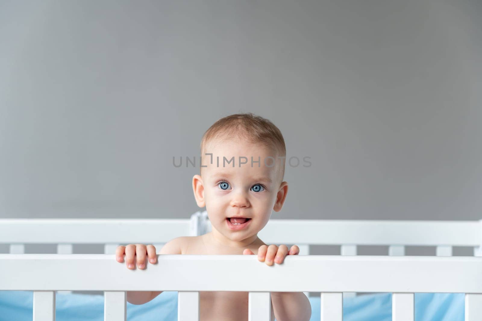 A modestly smiling baby leaned on the back of a wooden crib.