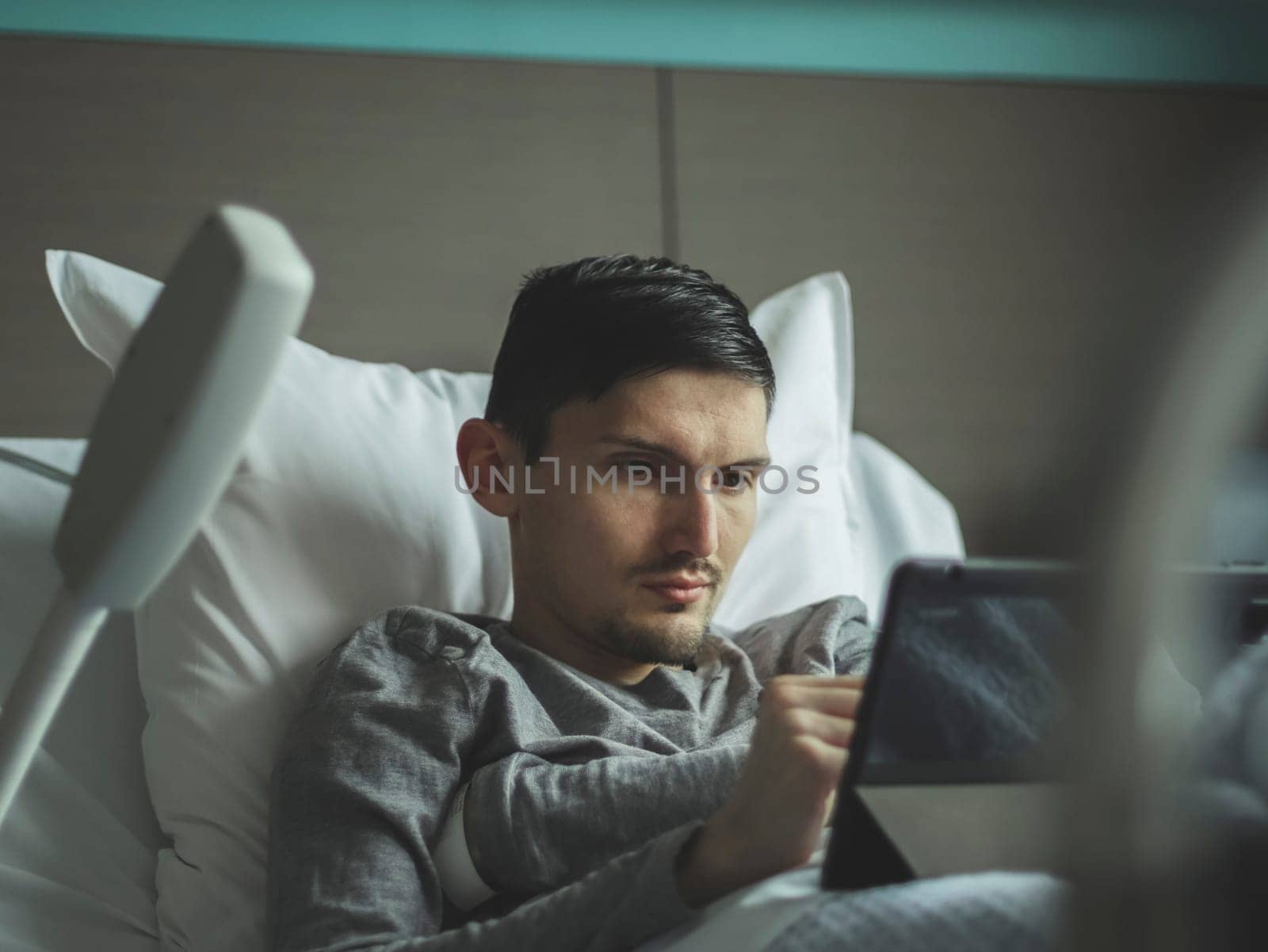 Portrait of one young caucasian guy lying in bed with watching a movie in a tablet in a hospital room, close-up side view.