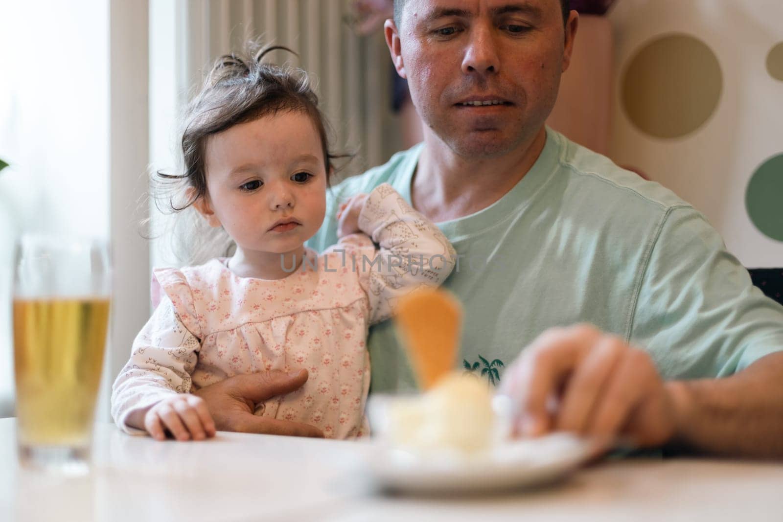Portrait of one handsome caucasian man with a little girl in her arms, sitting at a table with a drink in a glass and reaching for a dessert while sitting on a sofa in a cafe against the background of an abstract wall, close-up side view.