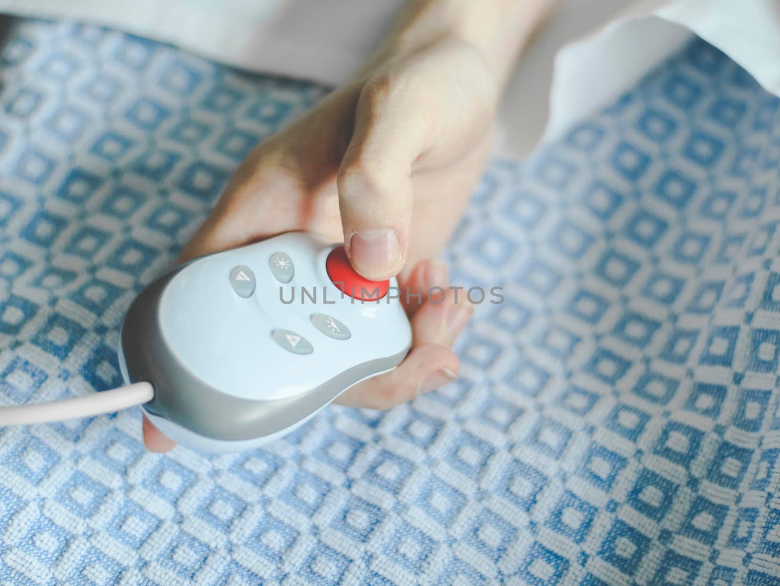 Hand of a young caucasian male patient lying in bed and holding a remote control in his hands, pressing a red button to call a nurse, close-up side view. Medical equipment concept.