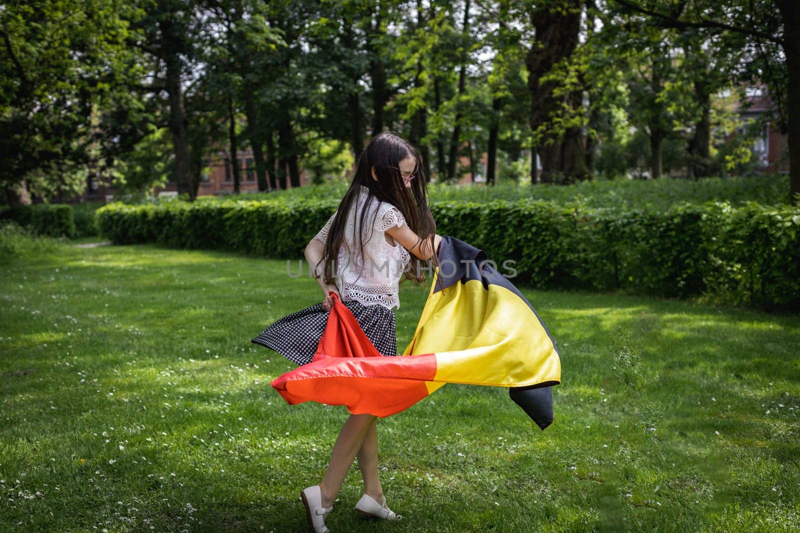 Portrait of one beautiful Caucasian little brunette girl with flowing hair twirling with a Belgian flag in a city park on a summer day, close-up side view.