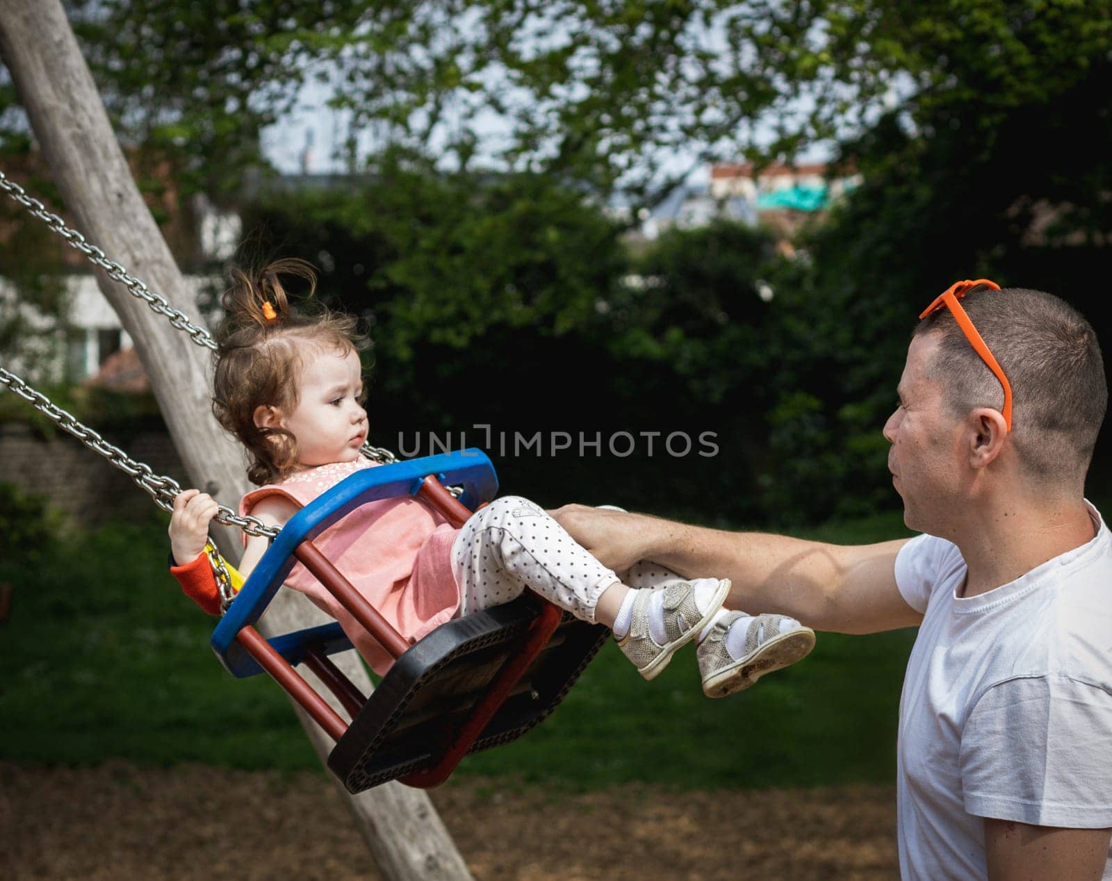 One handsome young Caucasian man father rides his little baby girl daughter on a swing on a summer day in the park on the playground, side view close-up.