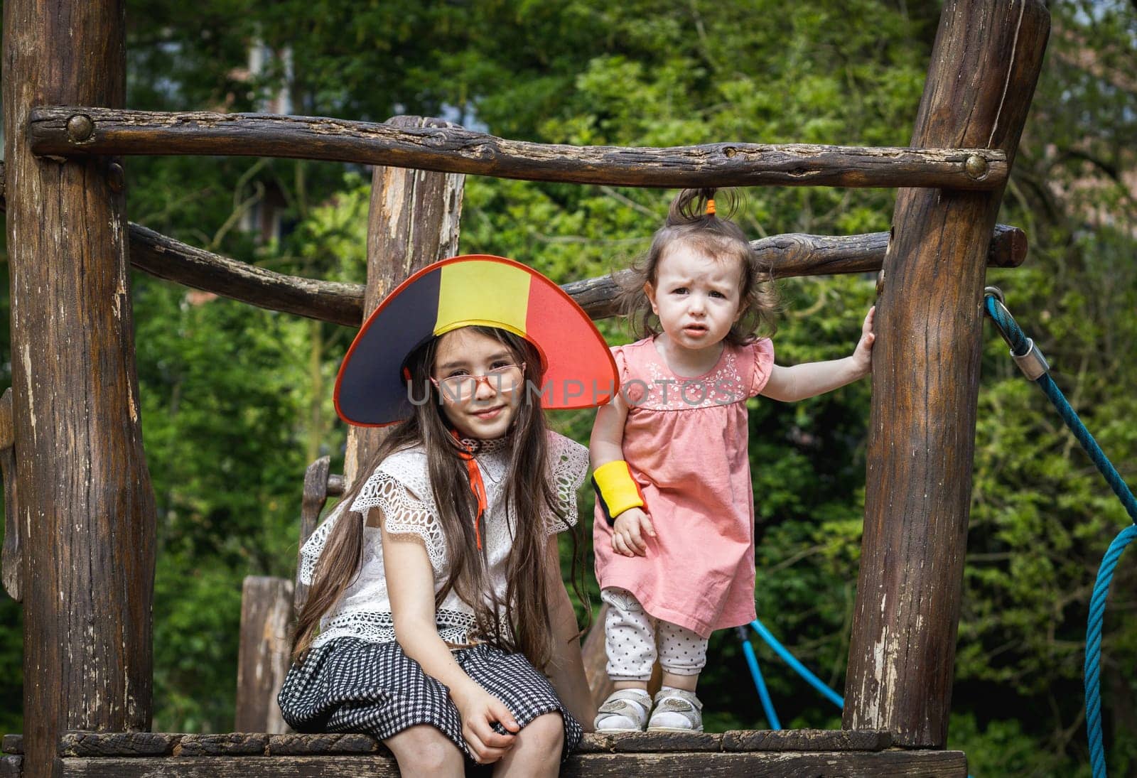 Portrait of two beautiful Caucasian little brunette girls in a Belgian flag hat sitting on a wooden walkway of a rope swing in a city park on a summer day, close-up side view.