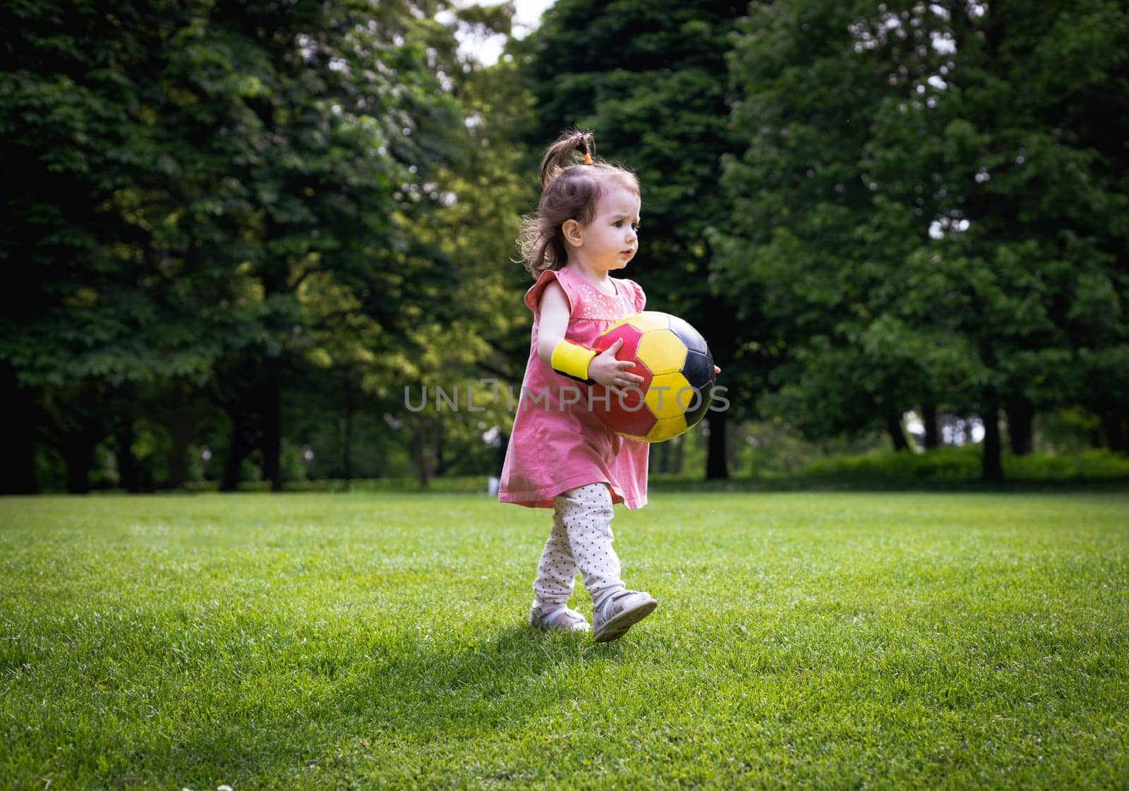 A little Caucasian girl in a dress is holding a soccer ball while running on the green lawn of a city park on a summer day, close-up view from the side.