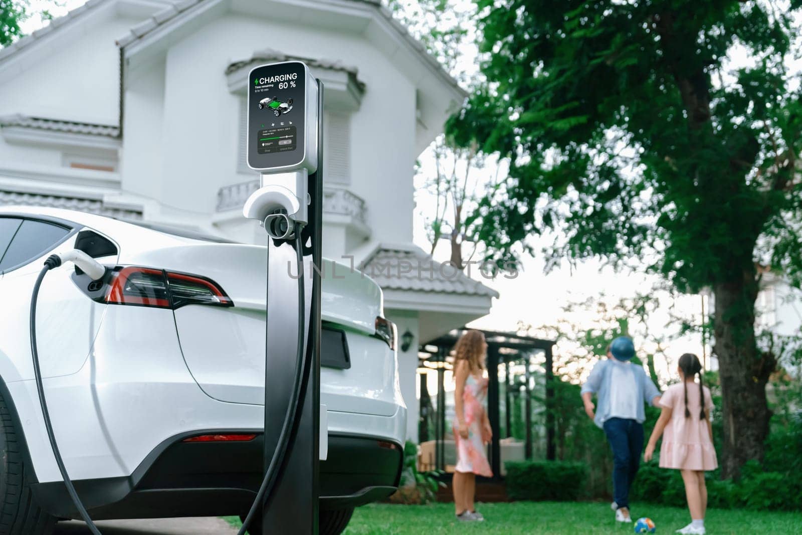 Focus electric vehicle recharge from home charging station on blur background of happy and playful family playing together. EV car using alternative and sustainable energy for better future.Synchronos