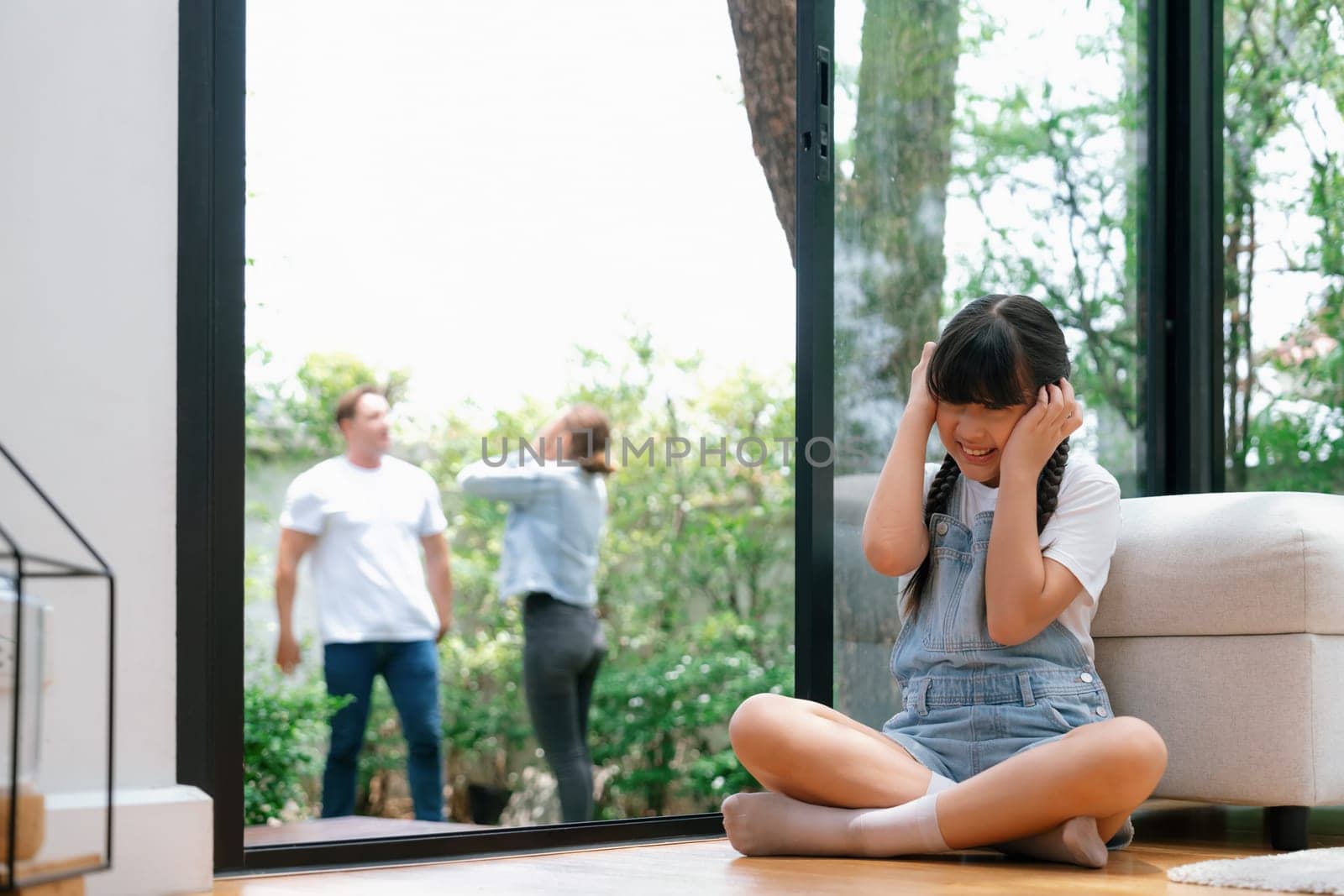 Stressed and unhappy girl huddle in corner, cover her ears with painful expression while her parent arguing in background. Domestic violence and traumatic childhood develop to depression. Synchronos