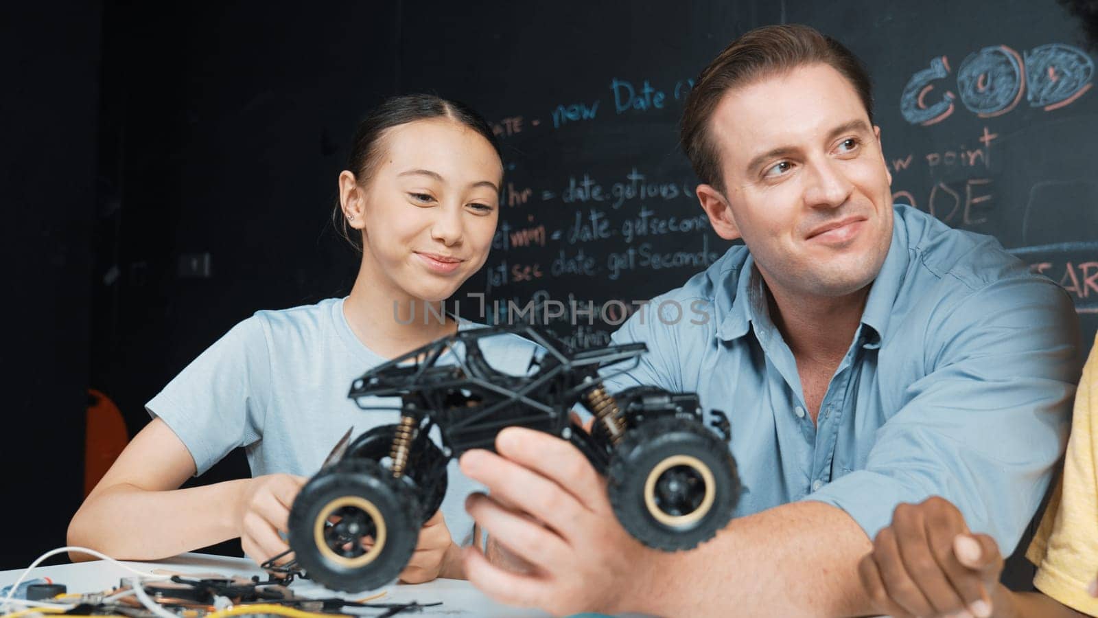 Smart teacher inspect car model construction to diverse student. Highschool children with mixed races listening mentor explain robotic model system at table with laptop and wires placed. Edification.