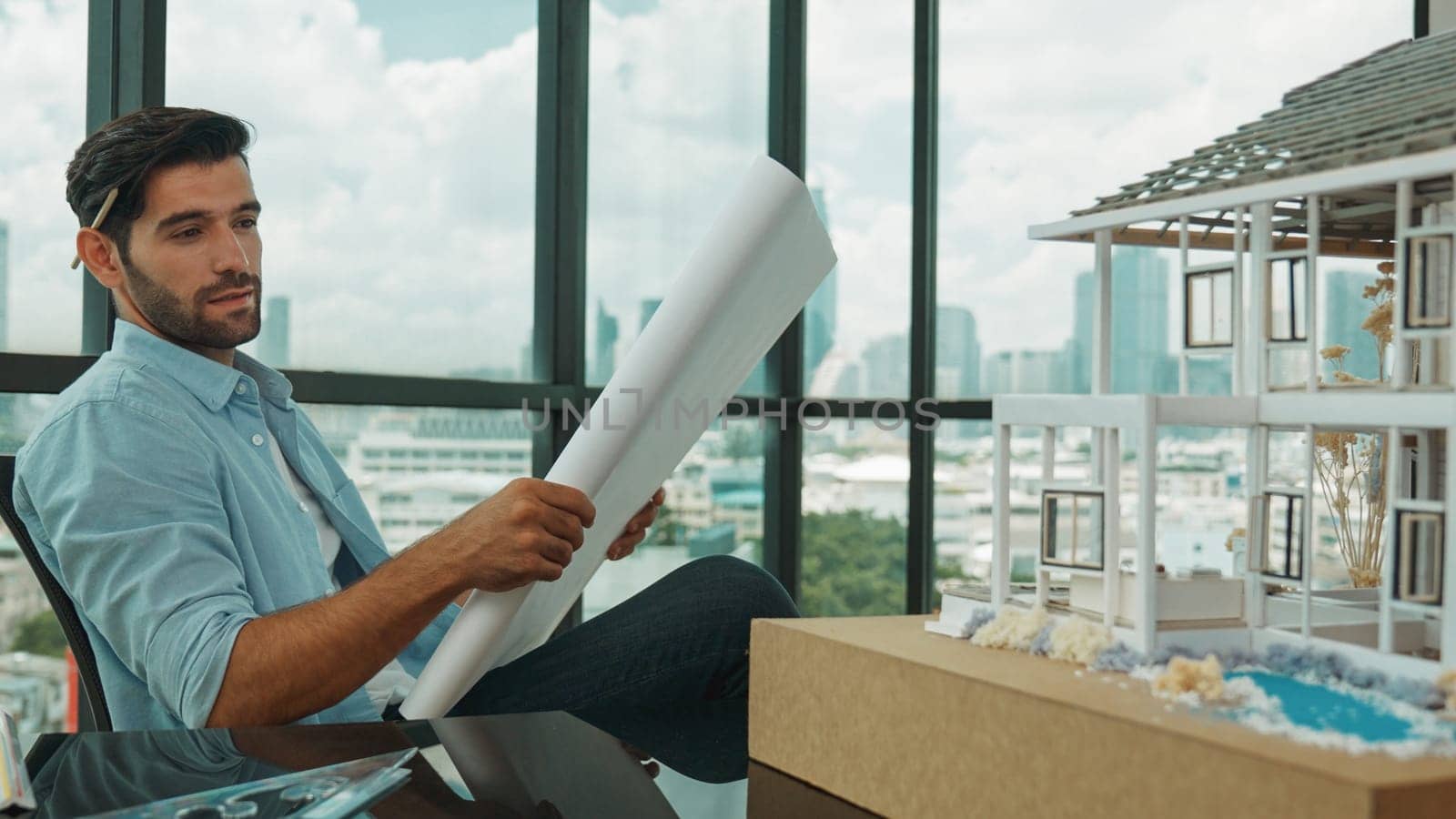 Smiling engineer looking project plan while design house model at office. Smart caucasian sitting relaxing while reading project plan. Creative design, civil engineering. Skyscraper. Tracery